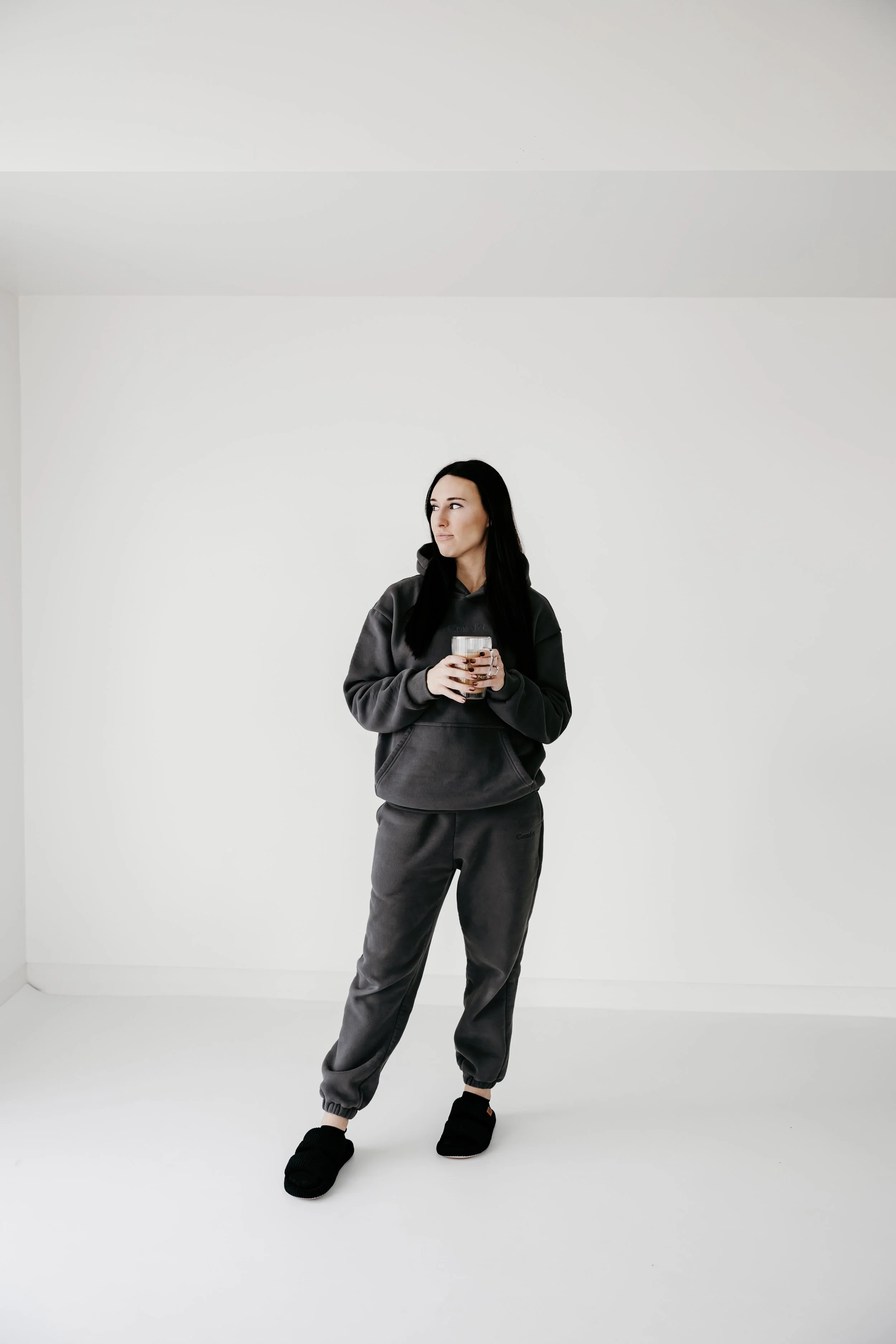 A woman in a gray hoodie and sweatpants holding a glass of coffee or tea in a sparse white room
