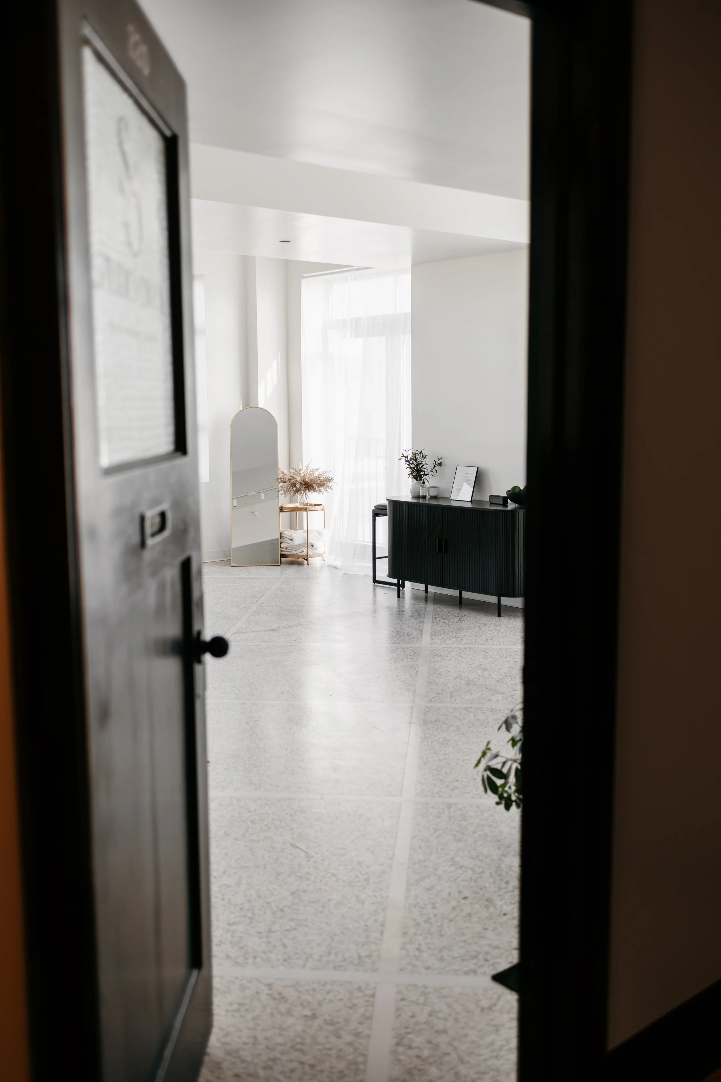 A minimalist, modern interior space visible through a partially open black door, featuring a light-colored speckled floor, a black sideboard with a plant and framed art, a white curved mirror, sheer white curtains, and natural light filling the room.