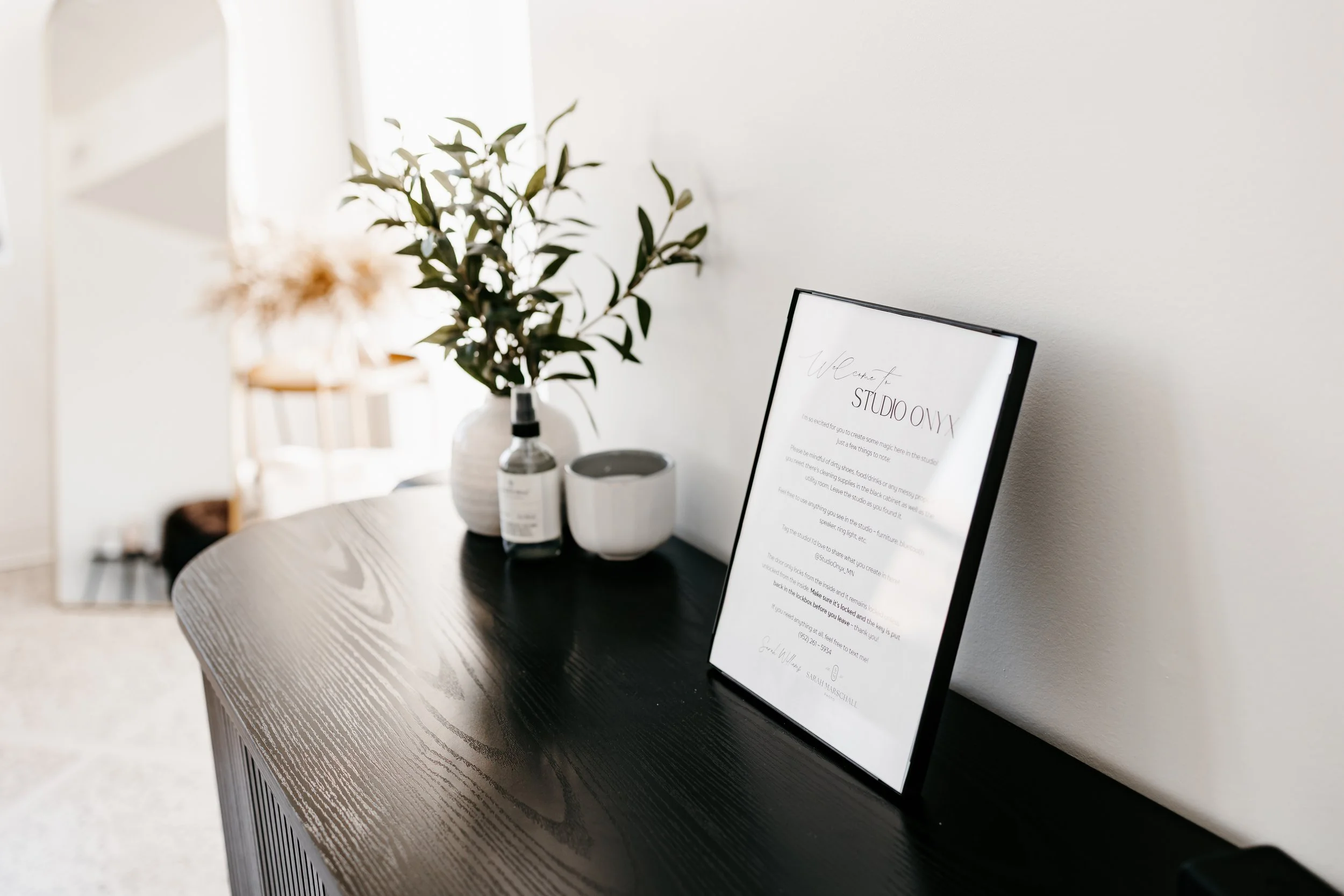 A black wooden console table with a framed sign, a potted plant, a diffuser, and a small bowl. The framed sign welcomes visitors to a studio.