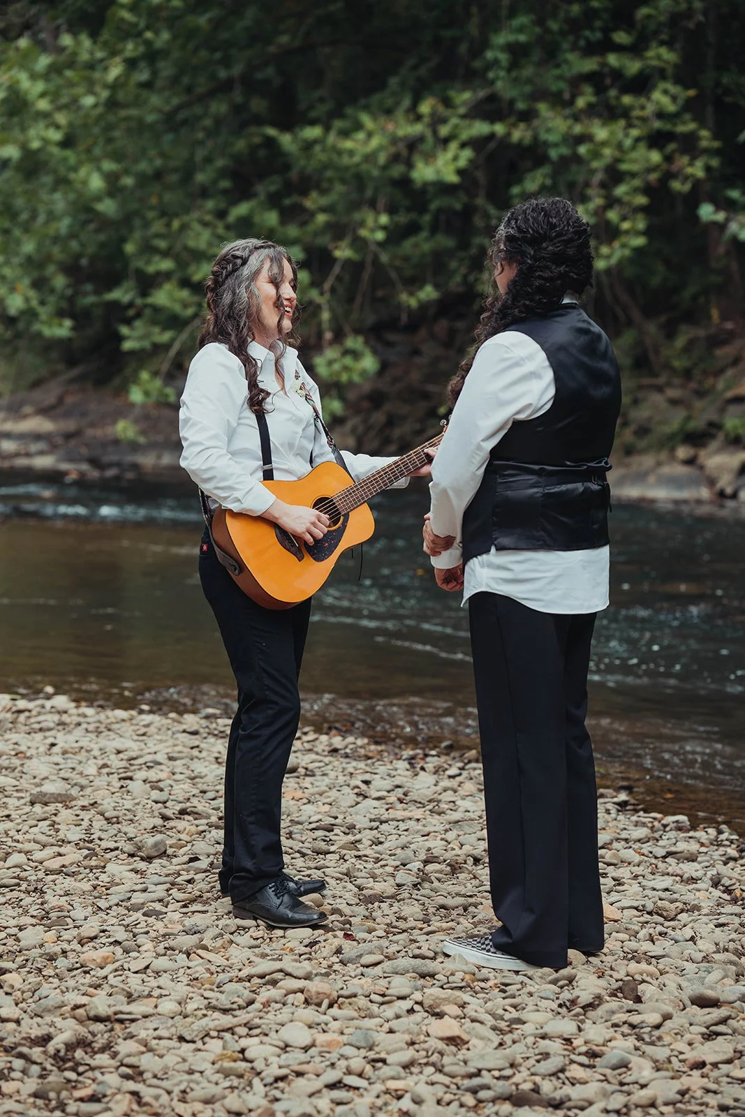 Two women standing on a rocky riverbank, one playing an acoustic guitar and the other listening, surrounded by trees.