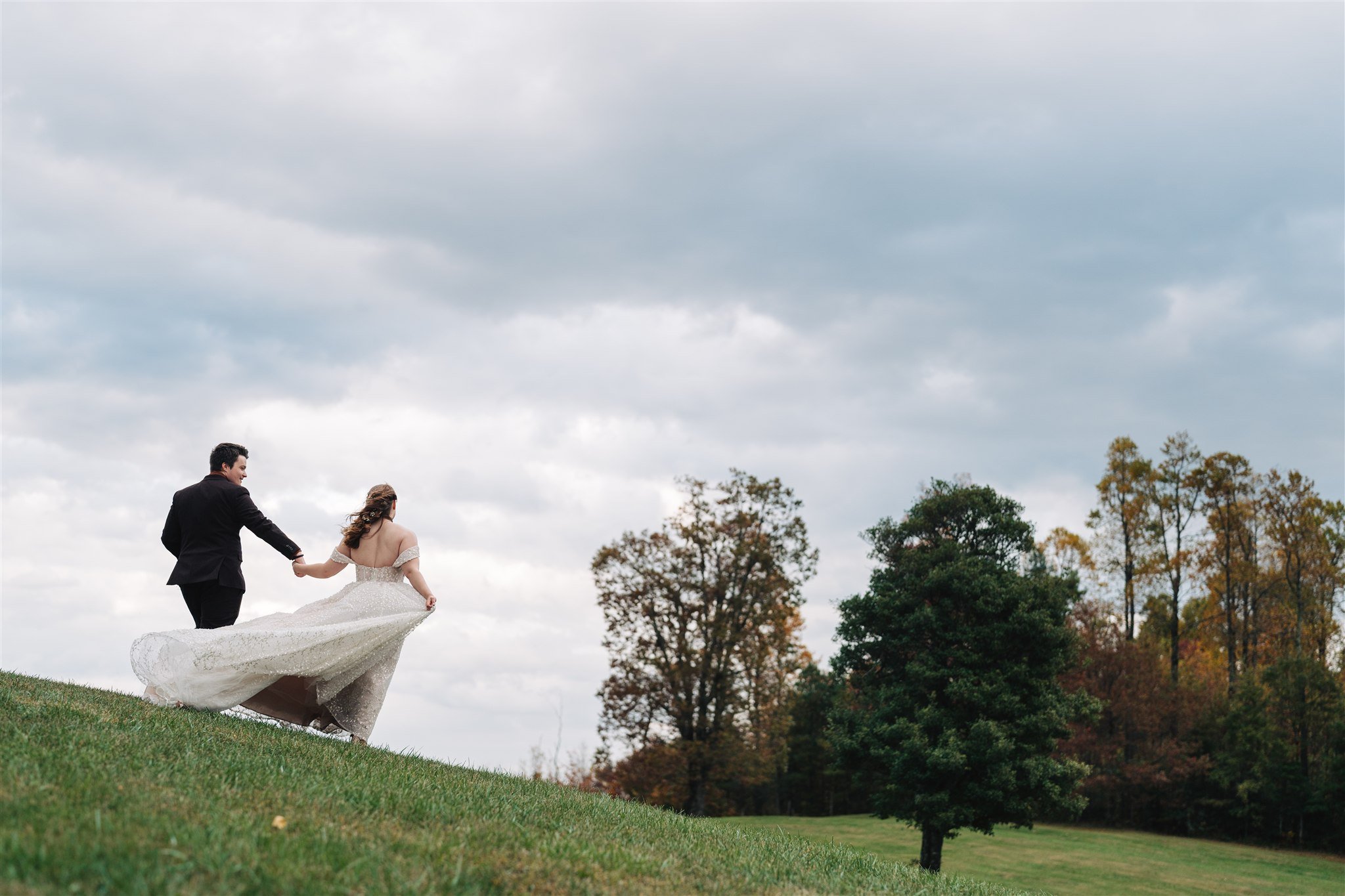 A bride and groom holding hands and walking on a grassy hill with trees and cloudy sky in the background.