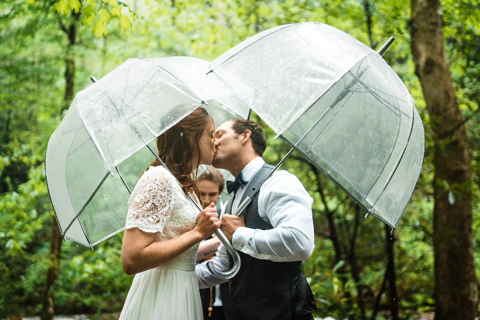 Bride and groom kissing under transparent umbrellas in a forest setting.