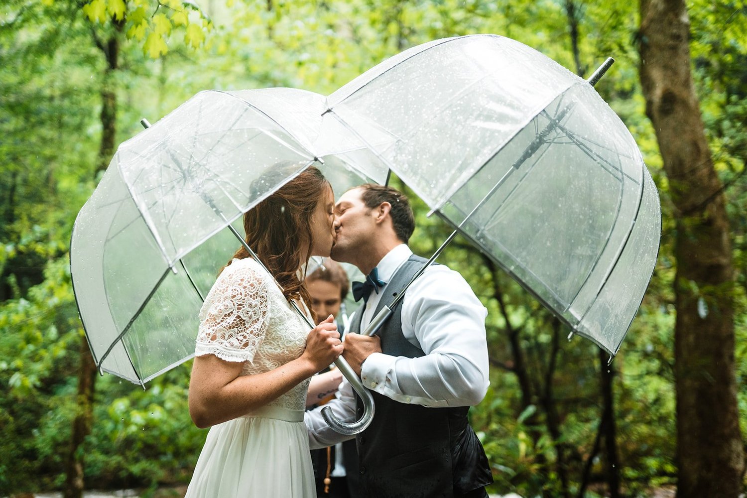 Wedding couple kissing beneath a clear umbrella during a rainy forest ceremony