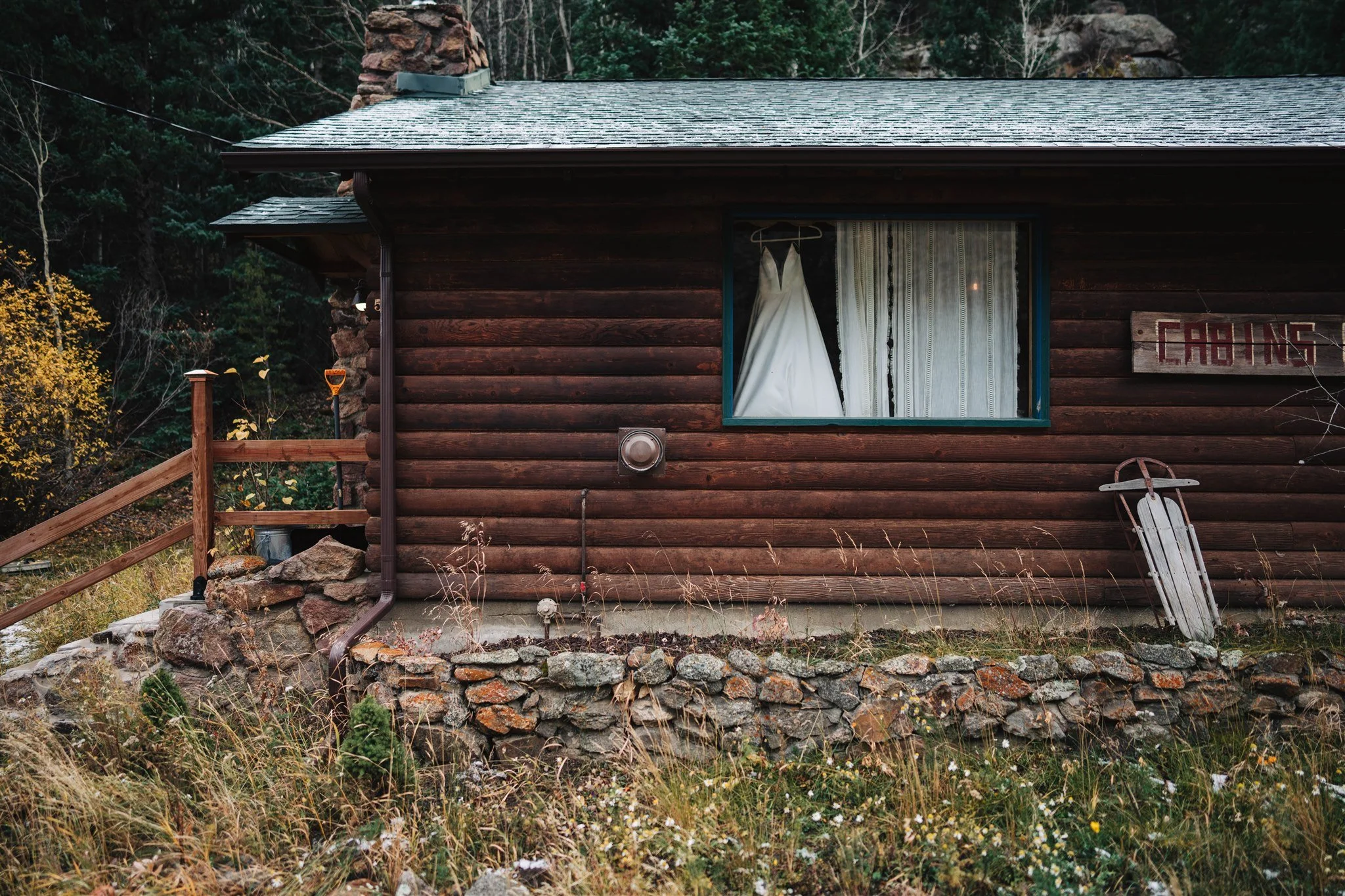 Rustic cabin with wood siding, a wedding dress hanging in the window, a sign reading 'CABINS,' and an old sled leaning against the wall.