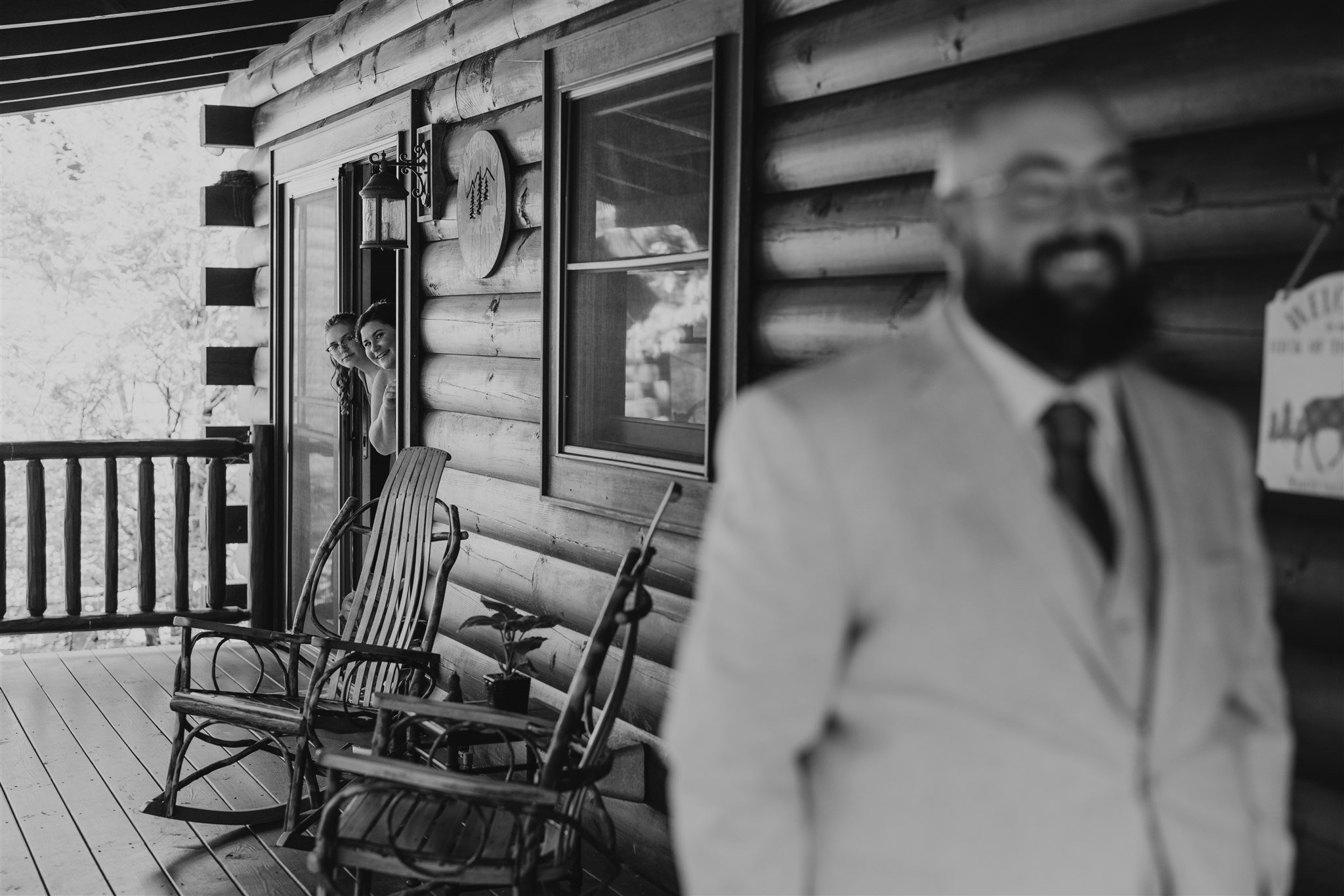 Two women peering out from a door window of a log cabin, smiling, with three rocking chairs on the porch, and a man standing in the foreground, out of focus. The setting appears to be outdoors with trees visible in the background.