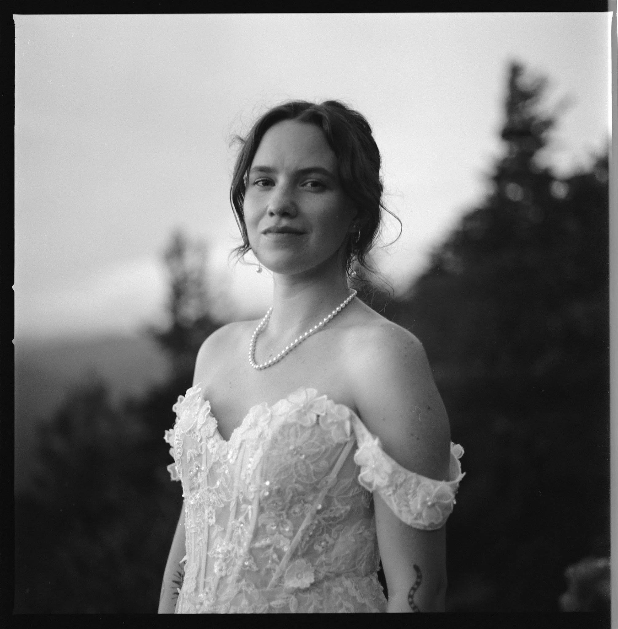 A black-and-white photo of a woman in an off-the-shoulder lace dress, wearing a string of pearls, outdoors with blurred trees in the background.