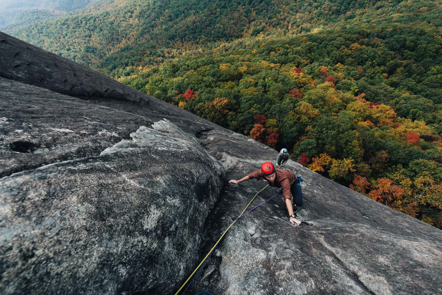 Two rock climbers ascending a mountain rock face above a colorful autumn forest