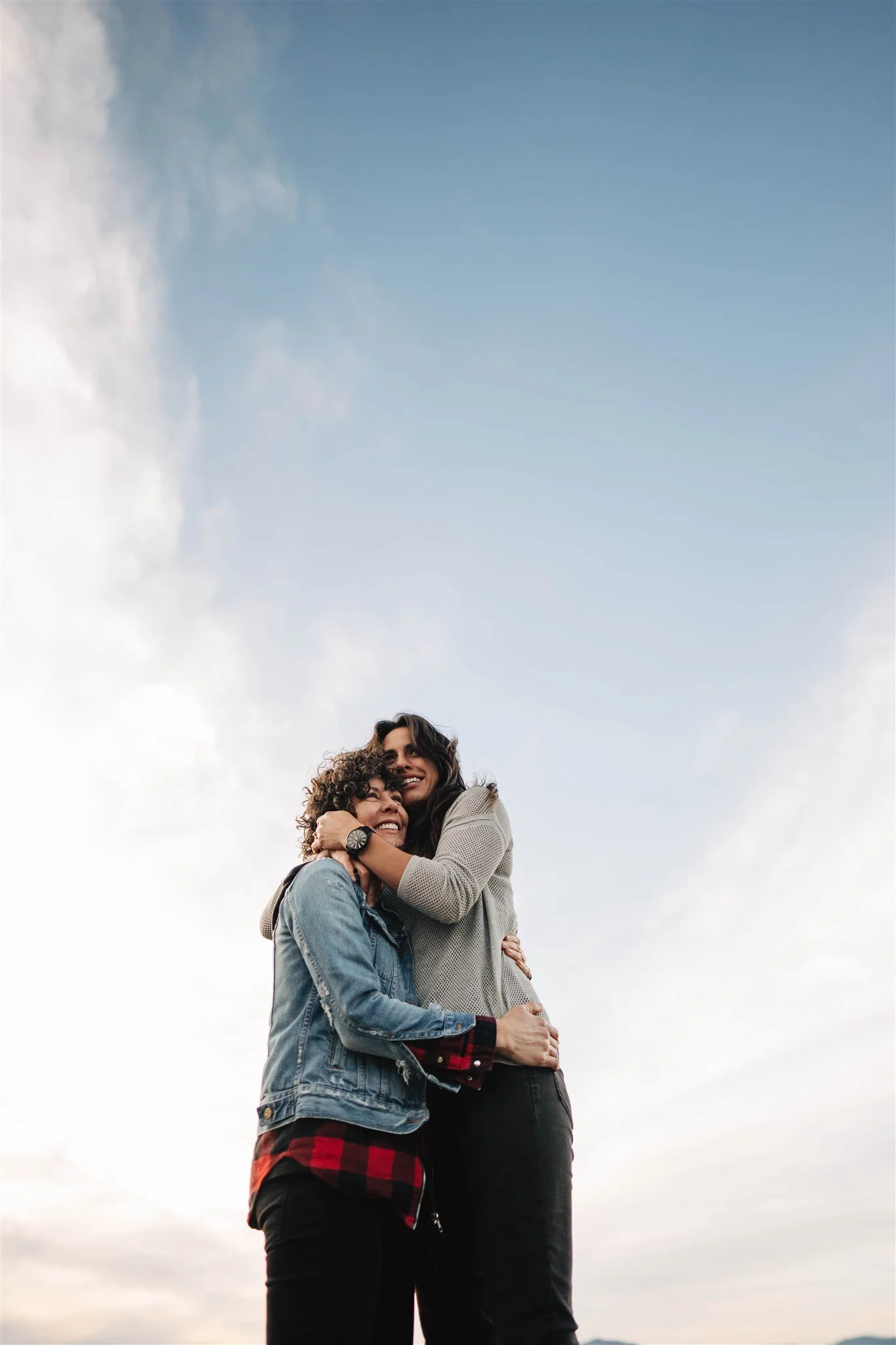 Two women hugging each other and smiling against a cloudy sky background.