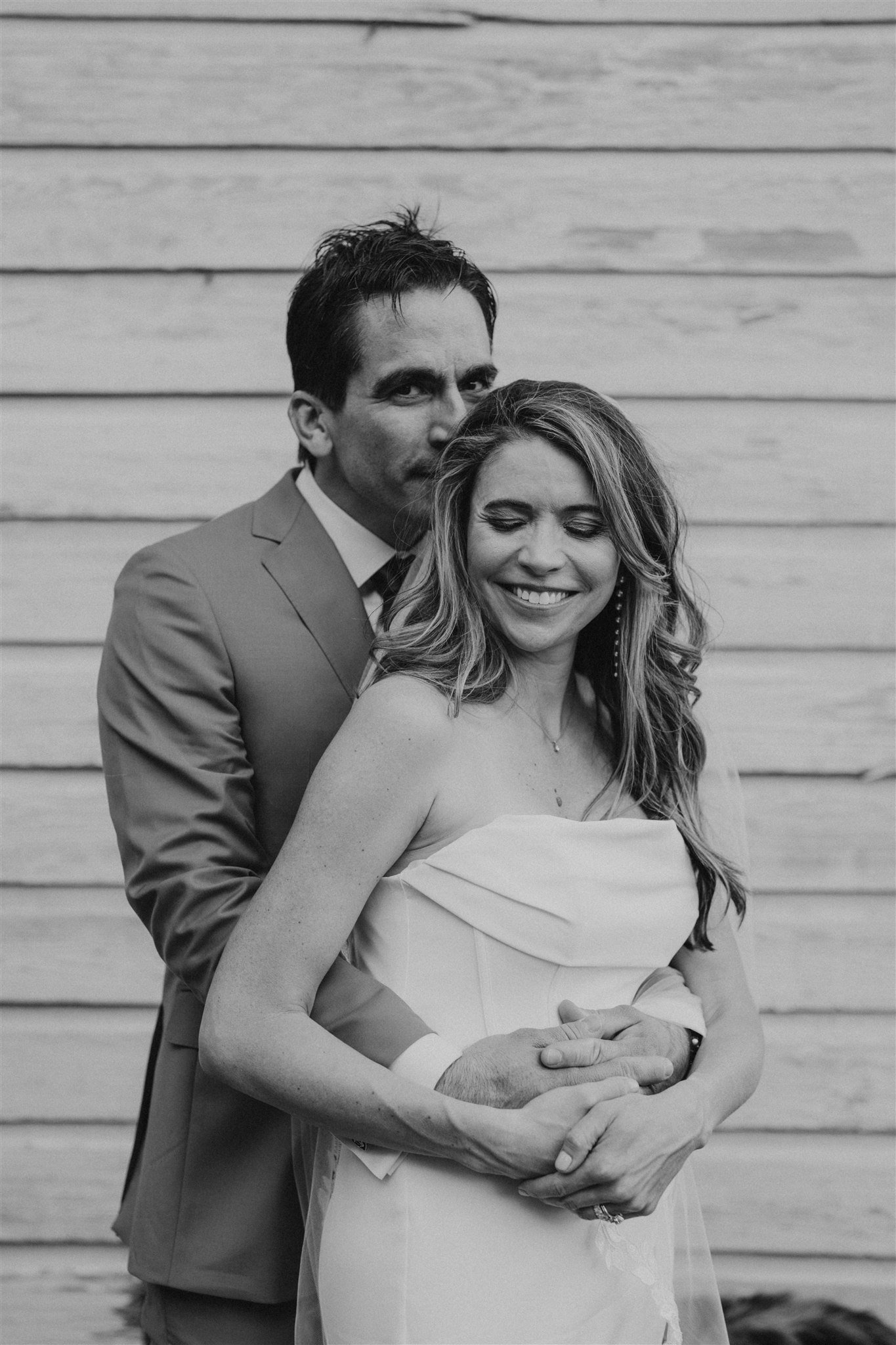 Black and white portrait of a couple sharing a quiet moment, with the bride smiling as the groom stands behind her.