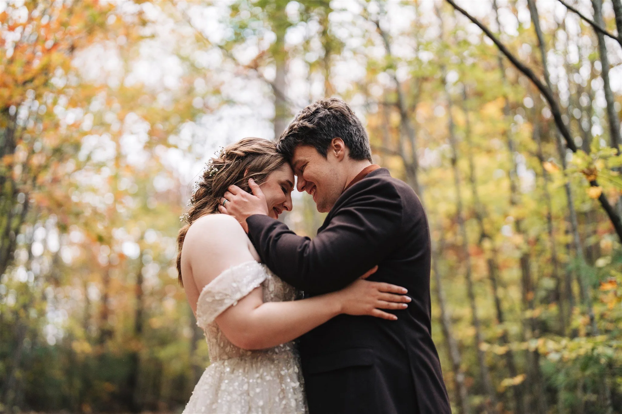 A couple embraces in a forest with autumn-colored leaves, smiling with foreheads touching.