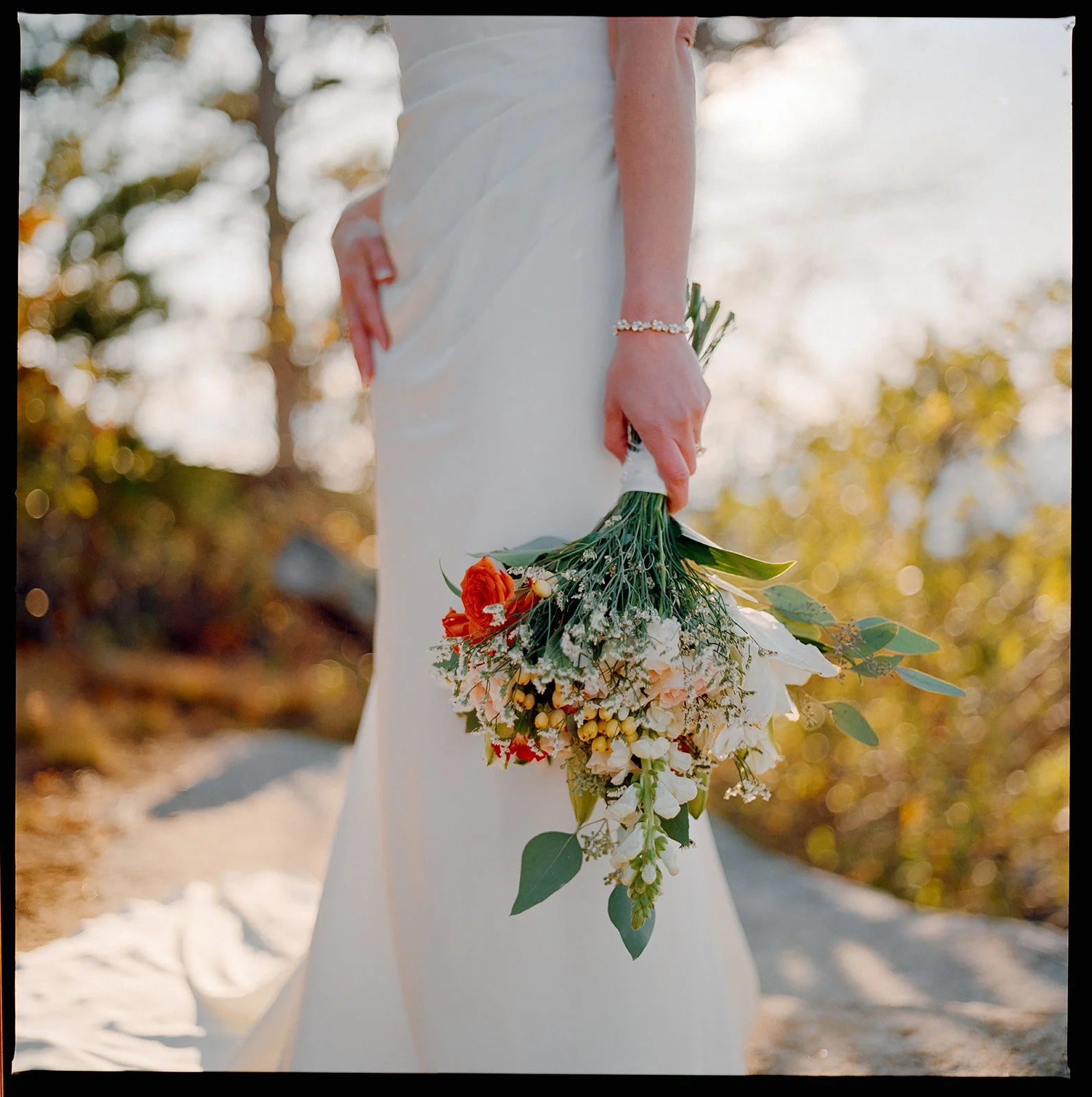 Bride in a white wedding dress holding a bouquet of flowers outdoors during sunset with trees and warm lighting in the background.