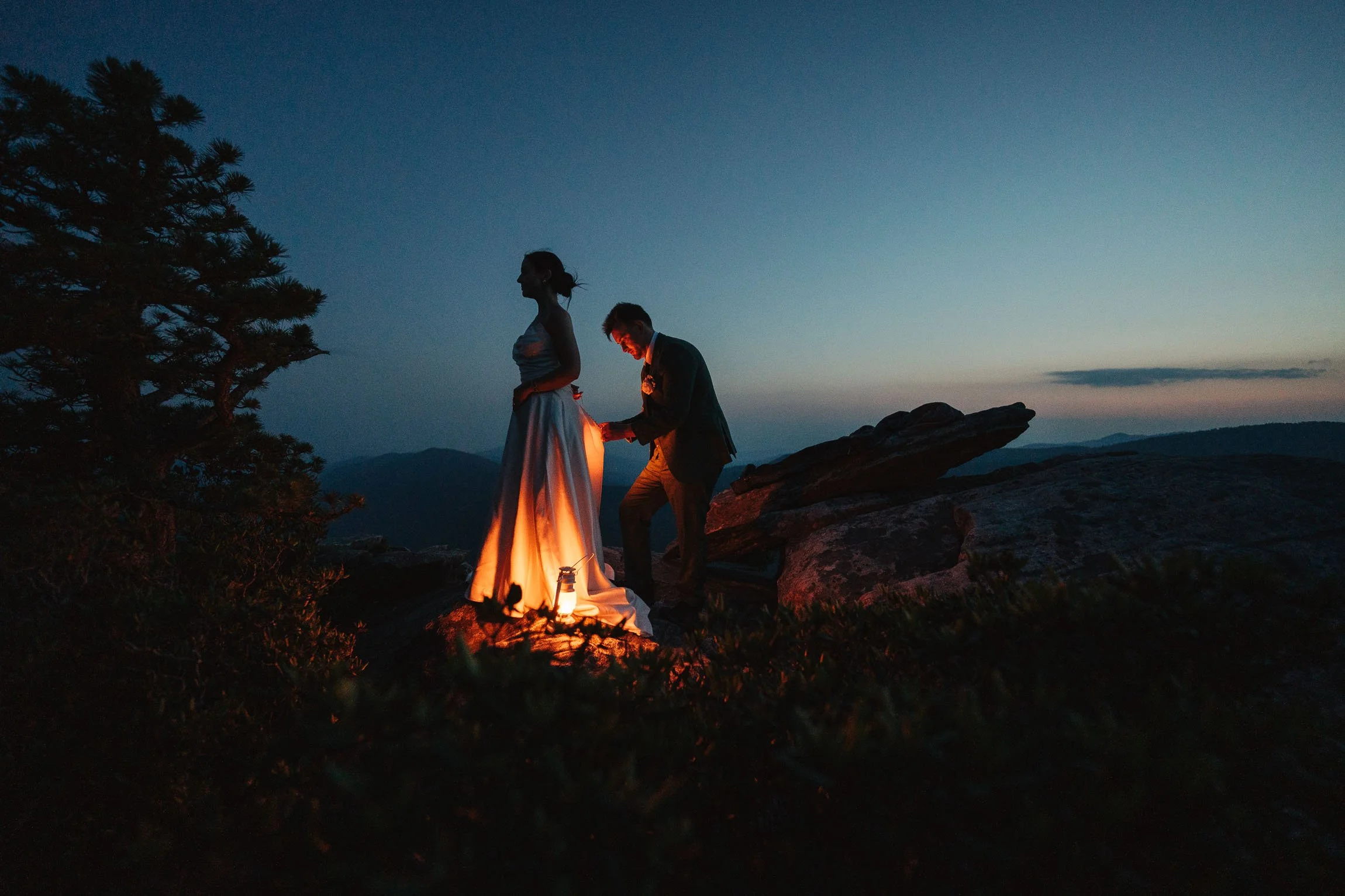 Bride and groom at blue hour on a rocky Linville Gorge summit, with the groom adjusting her dress as a small lantern casts warm light against the darkening mountain horizon.