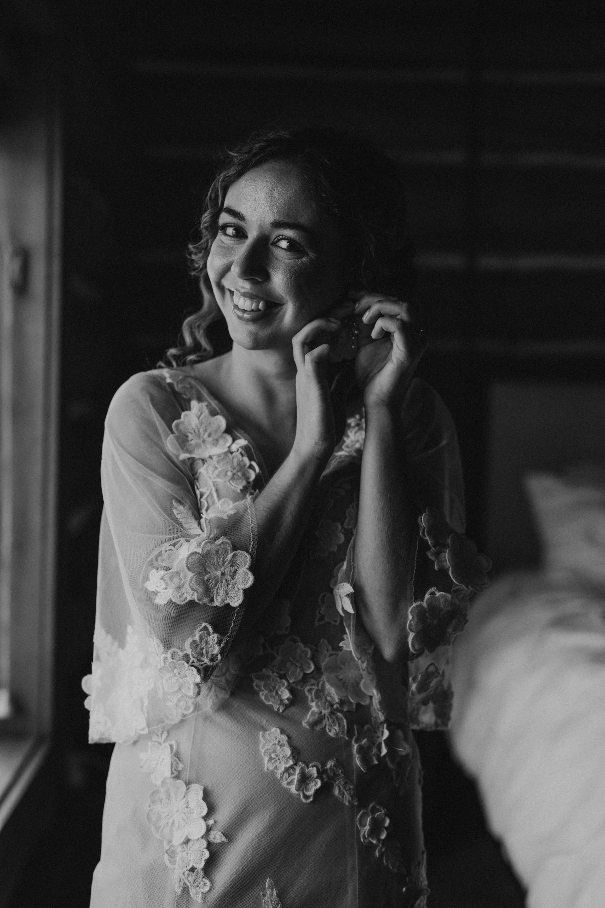 A woman smiling and putting on earrings in a room with natural light, wearing a floral embroidered sheer dress.
