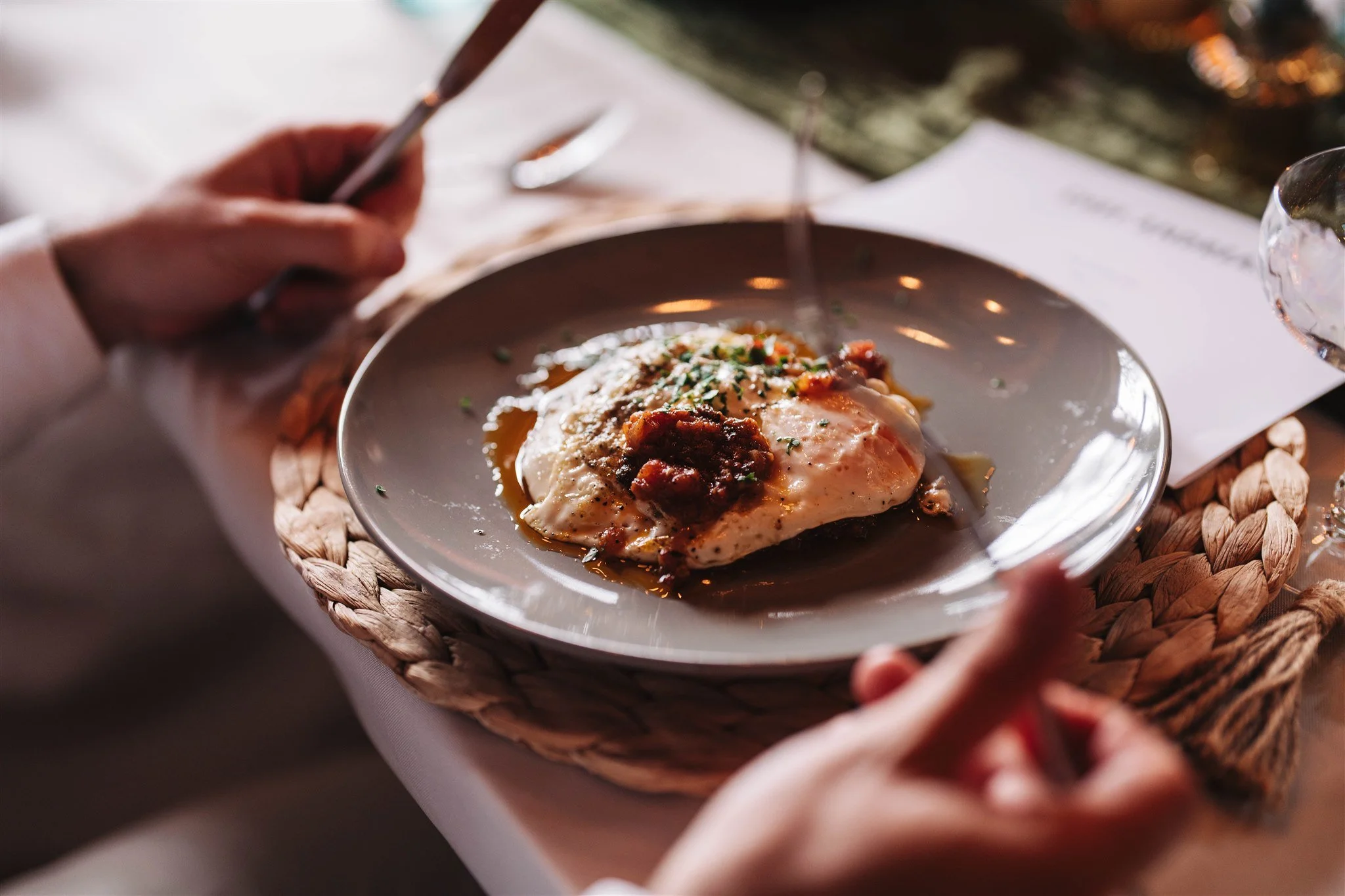 A person holding a knife and fork, about to eat a dish, possibly eggs, served on a gray plate with a textured placemat.