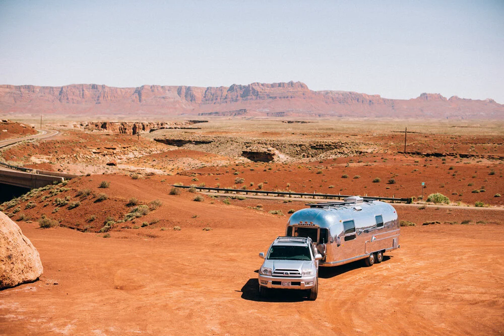 Car towing an Airstream trailer is parked on a dirt lot in a desert landscape with red rock formations and mountains in the background.