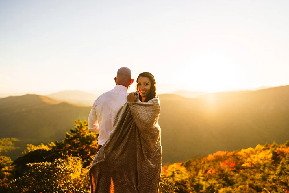 Bride wrapped in a warm blanket beside her groom during a fall mountain sunset