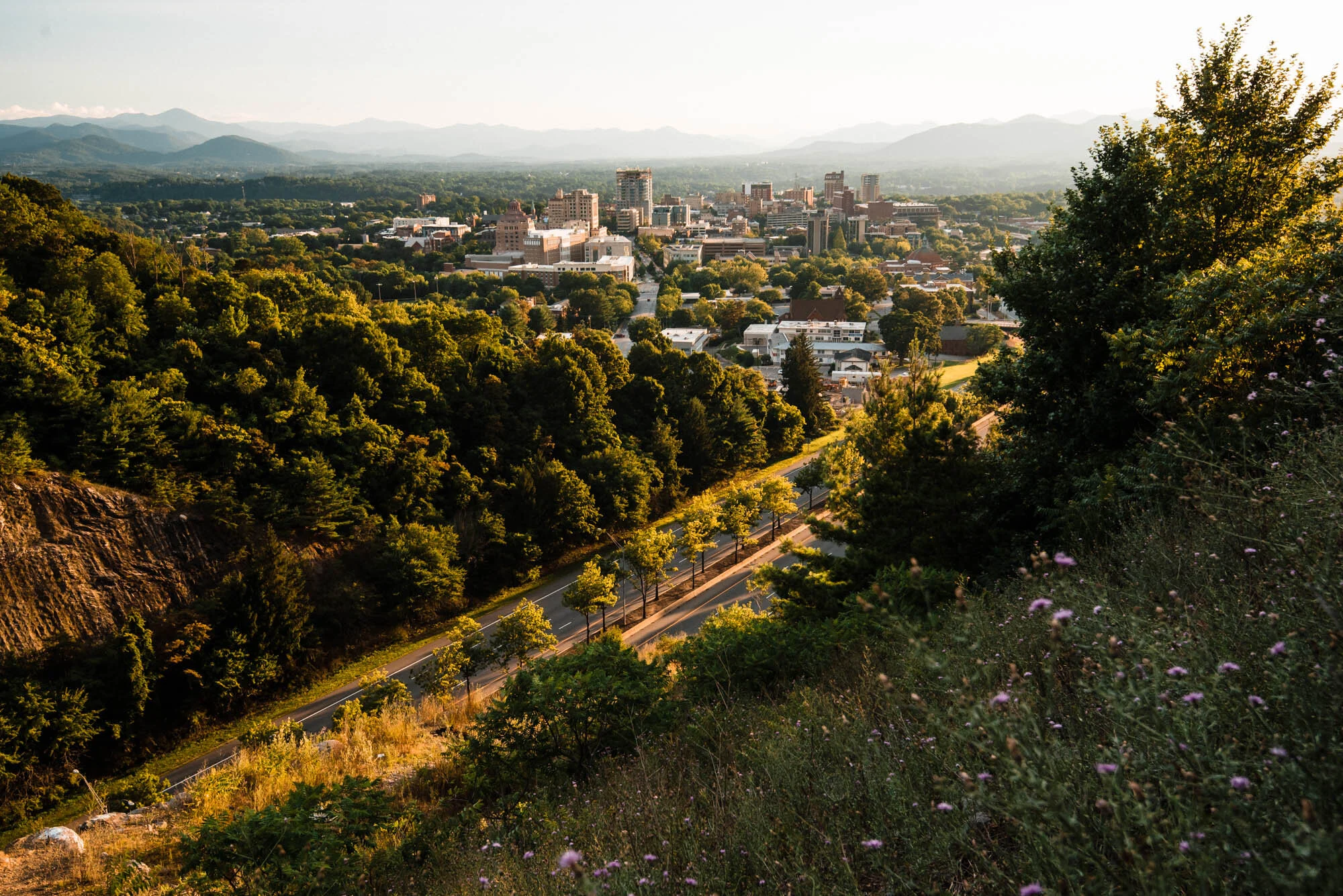 Aerial view of a city with tall buildings, surrounded by green trees and mountains in the background, during sunset.