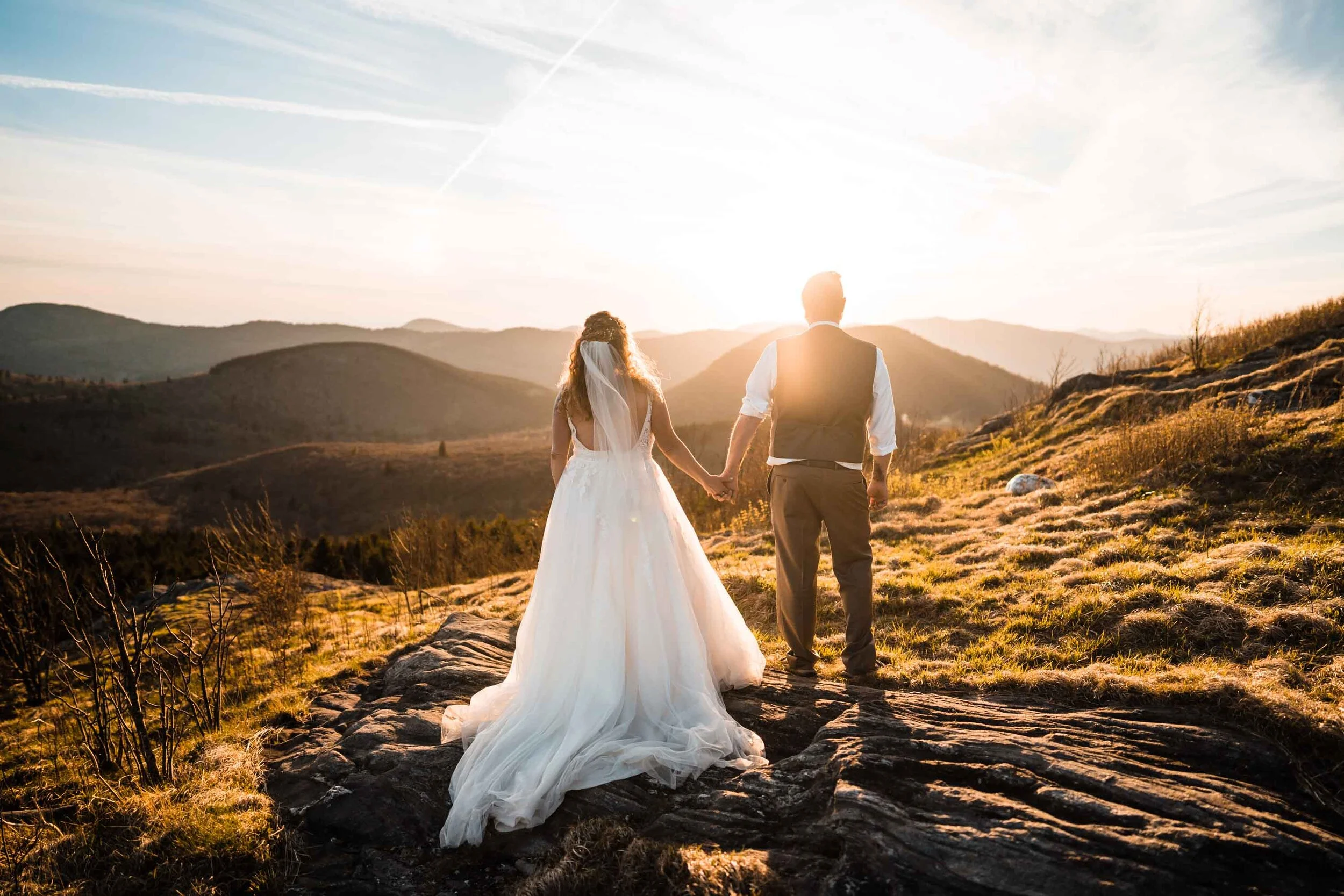 A bride and groom watch the sun go down on a mountaintop in their wedding clothes.