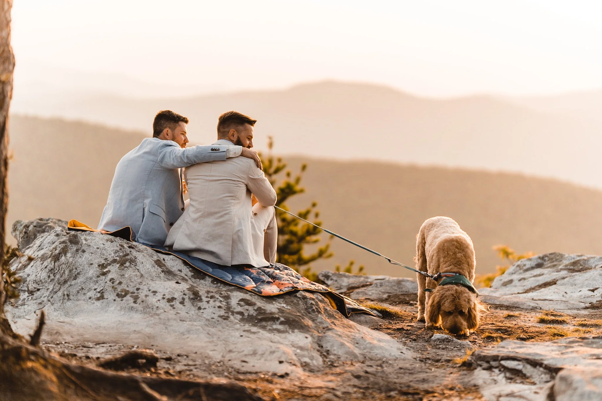 Two men sitting on a large rock with a blanket, overlooking mountains at sunset, with a dog on a leash exploring nearby.
