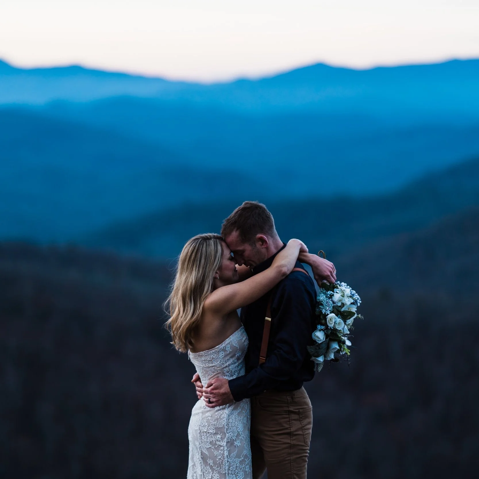A couple embracing, standing outdoors in front of a scenic mountain view at dusk. The woman is wearing a white dress and holding a bouquet of flowers.