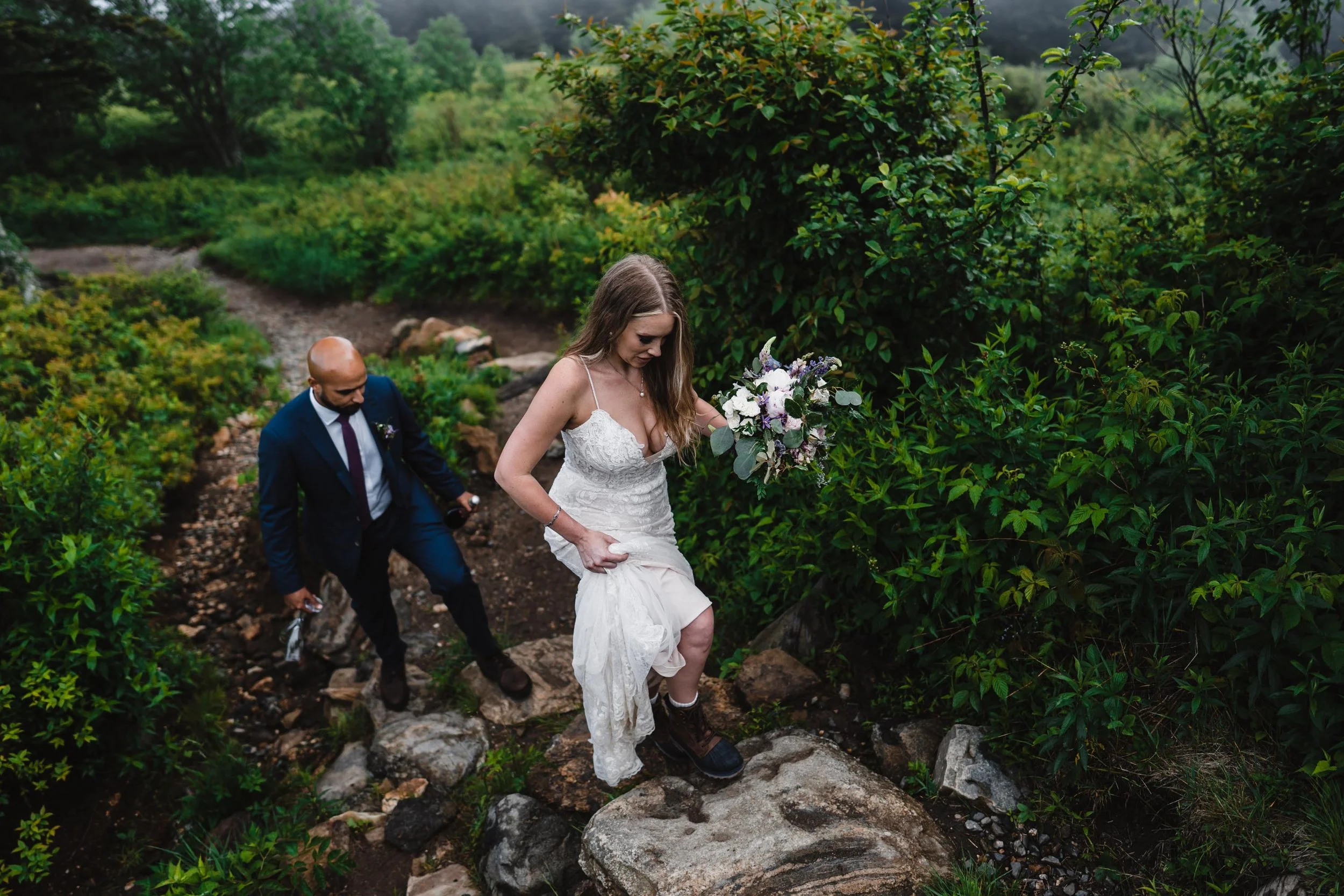A couple hikes through vivid green mountain laurel and other plants in their wedding clothes.