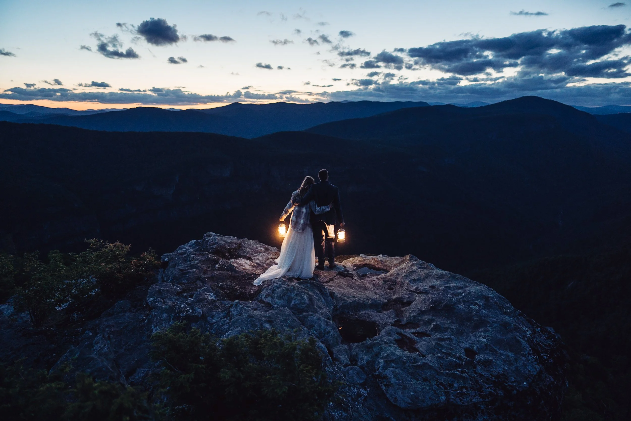A newly married couple hold each other while holding lanterns as night falls on a rocky ridge.