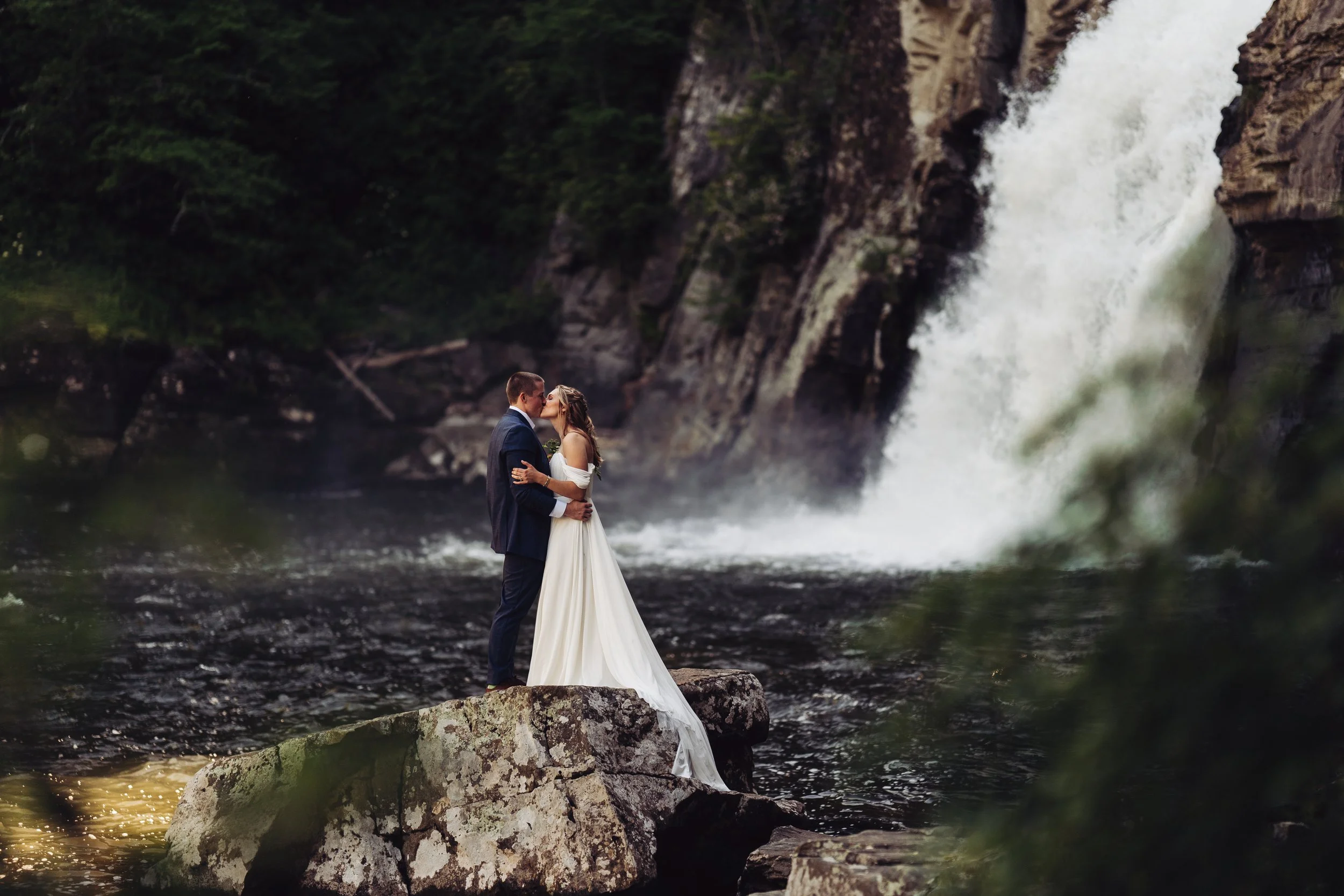 A couple embrace and kiss in front of a large waterfall in their wedding clothes.