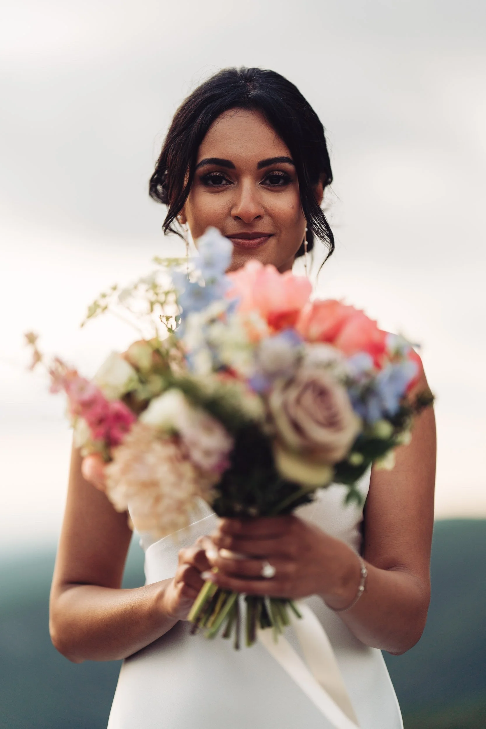 A woman in a white dress holding a bouquet of colorful flowers, standing outdoors with a cloudy sky in the background.