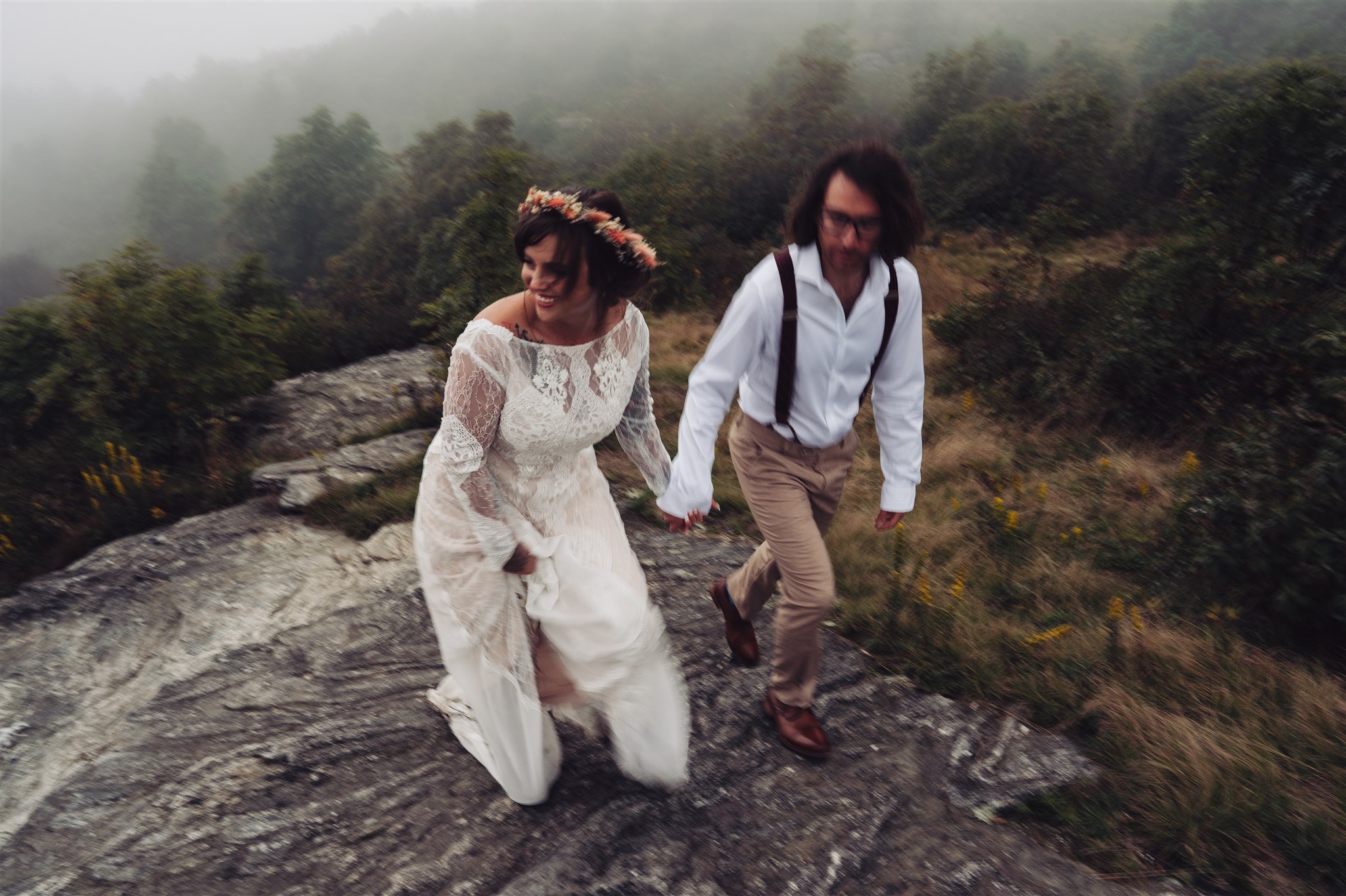 A woman in a white lace wedding dress and a floral crown holding hands with a man in a white shirt, khaki pants, and suspenders, walking on a rocky trail in a foggy, forested area.