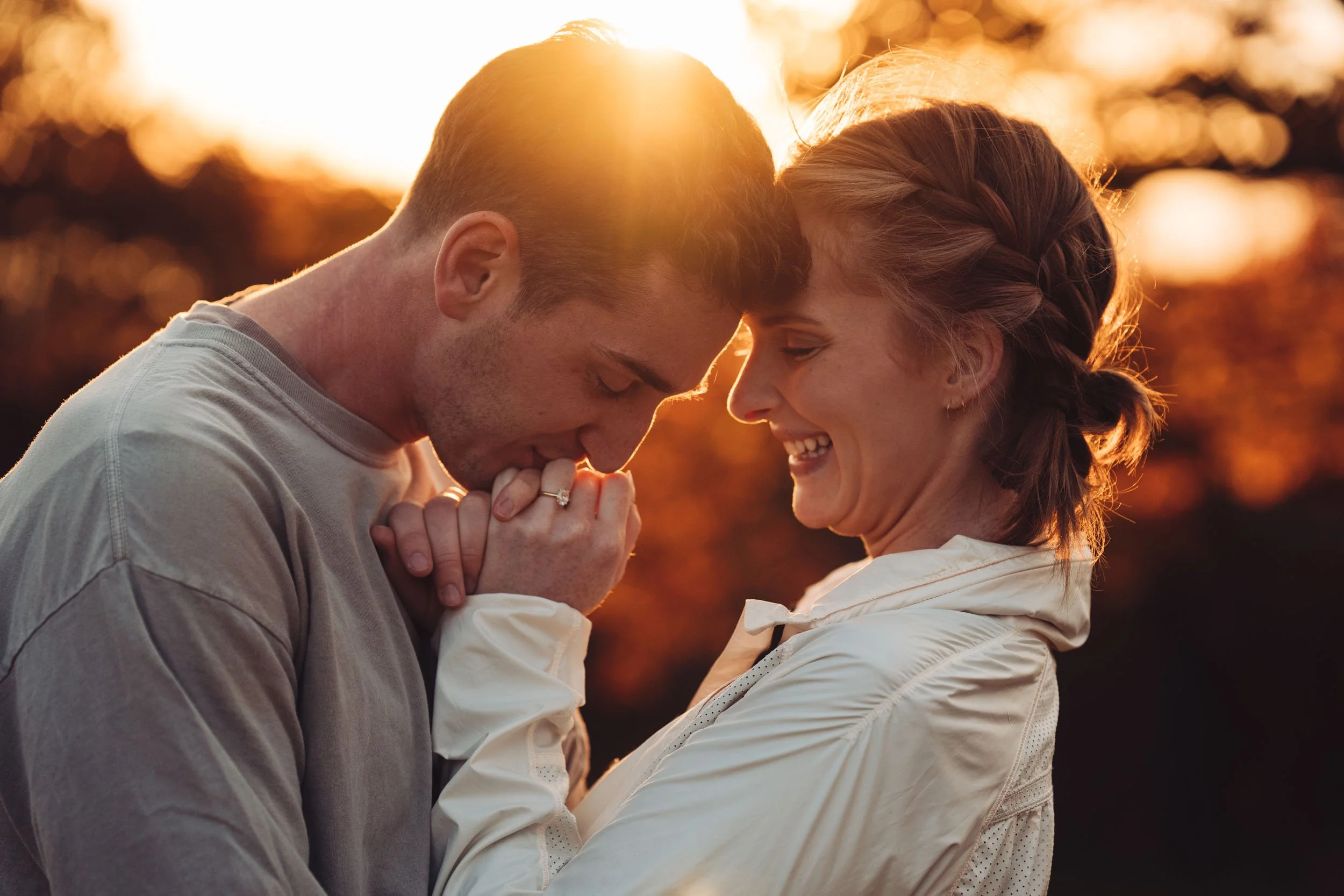 Couple holding hands while guy kisses his fiance's hand as the sun sets.