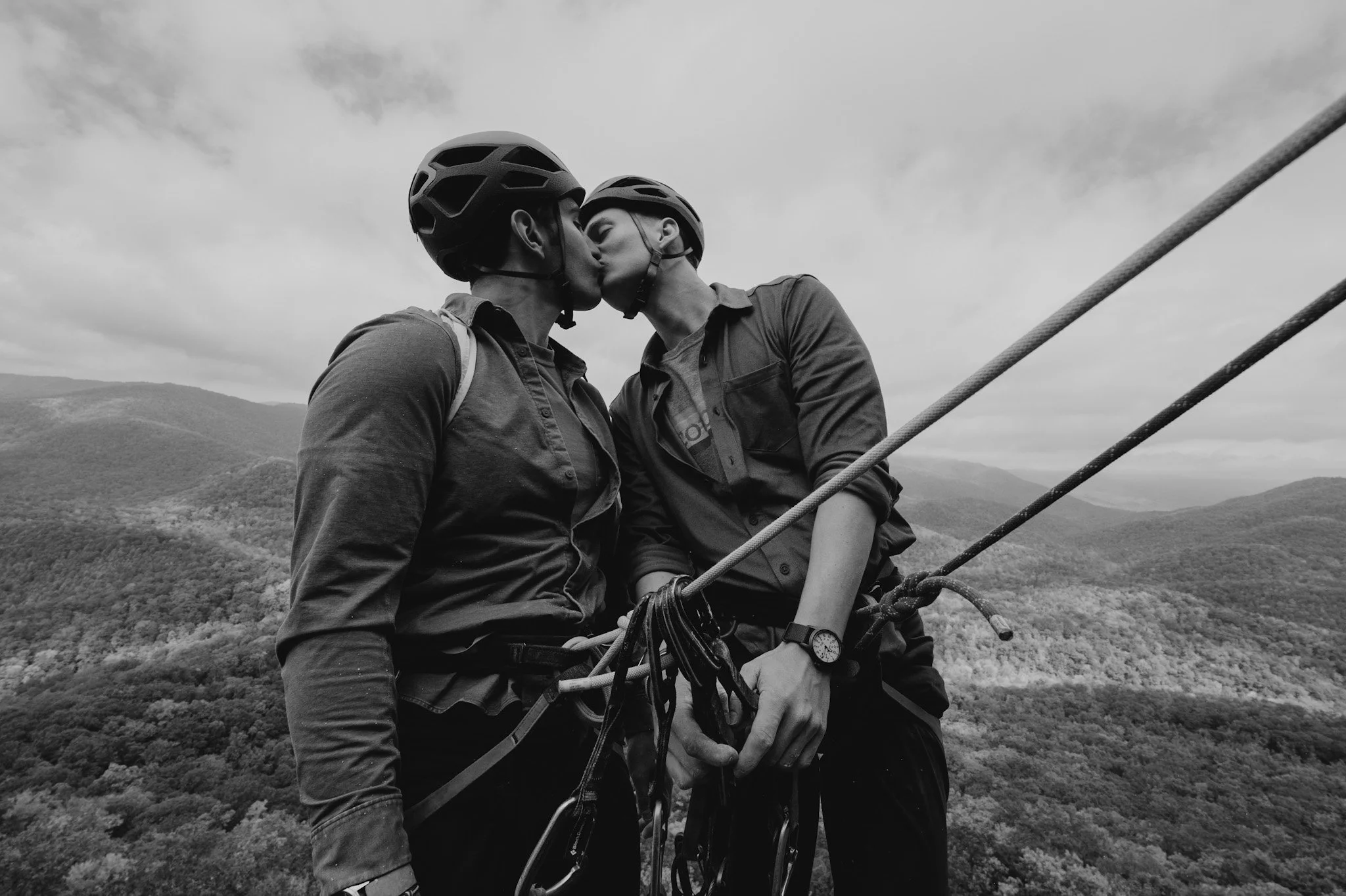 Two climbers in helmets kissing atop a mountain with a forested landscape in the background.