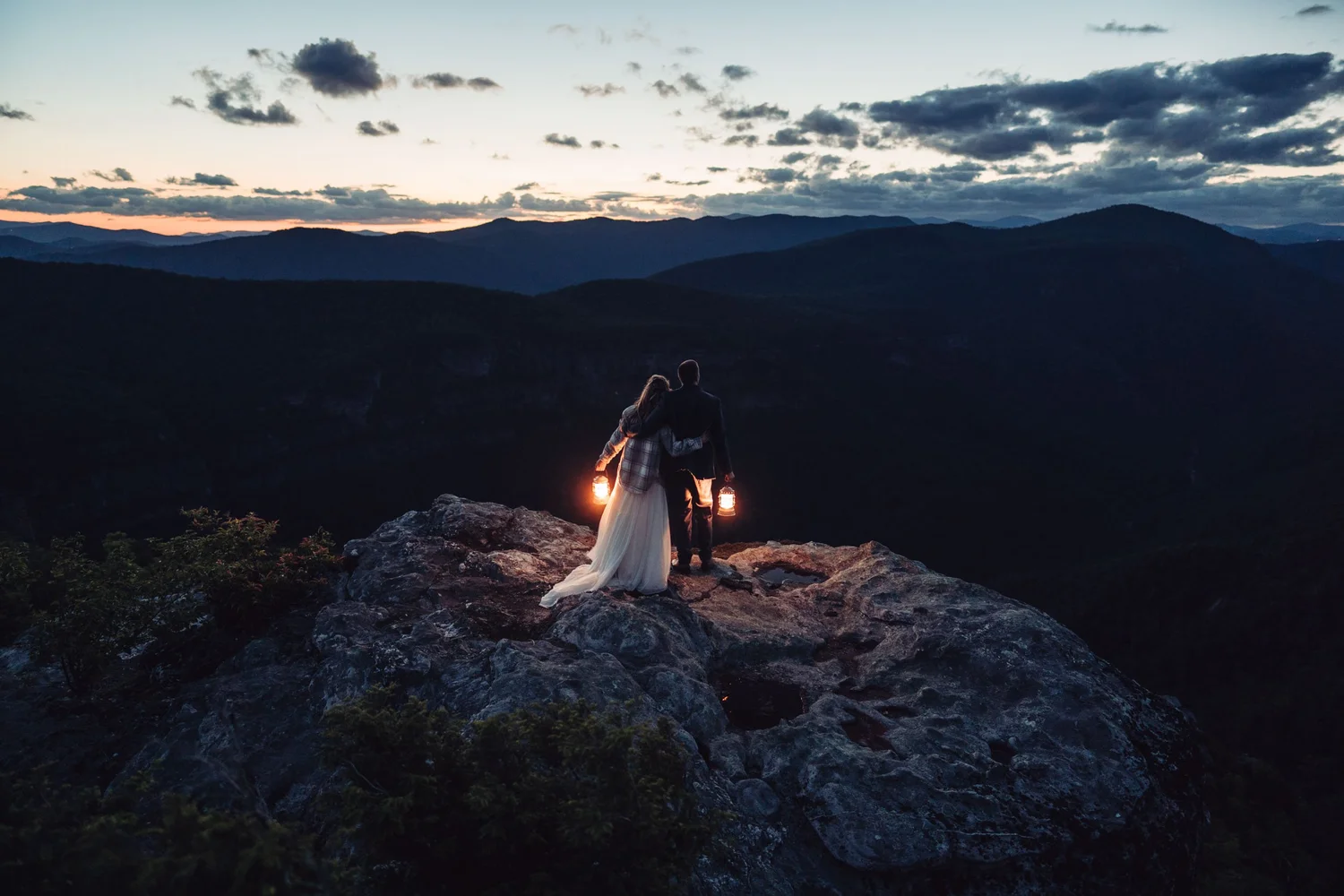 Kaitlyn and Evan standing together with glowing lanterns above Linville Gorge after sunset