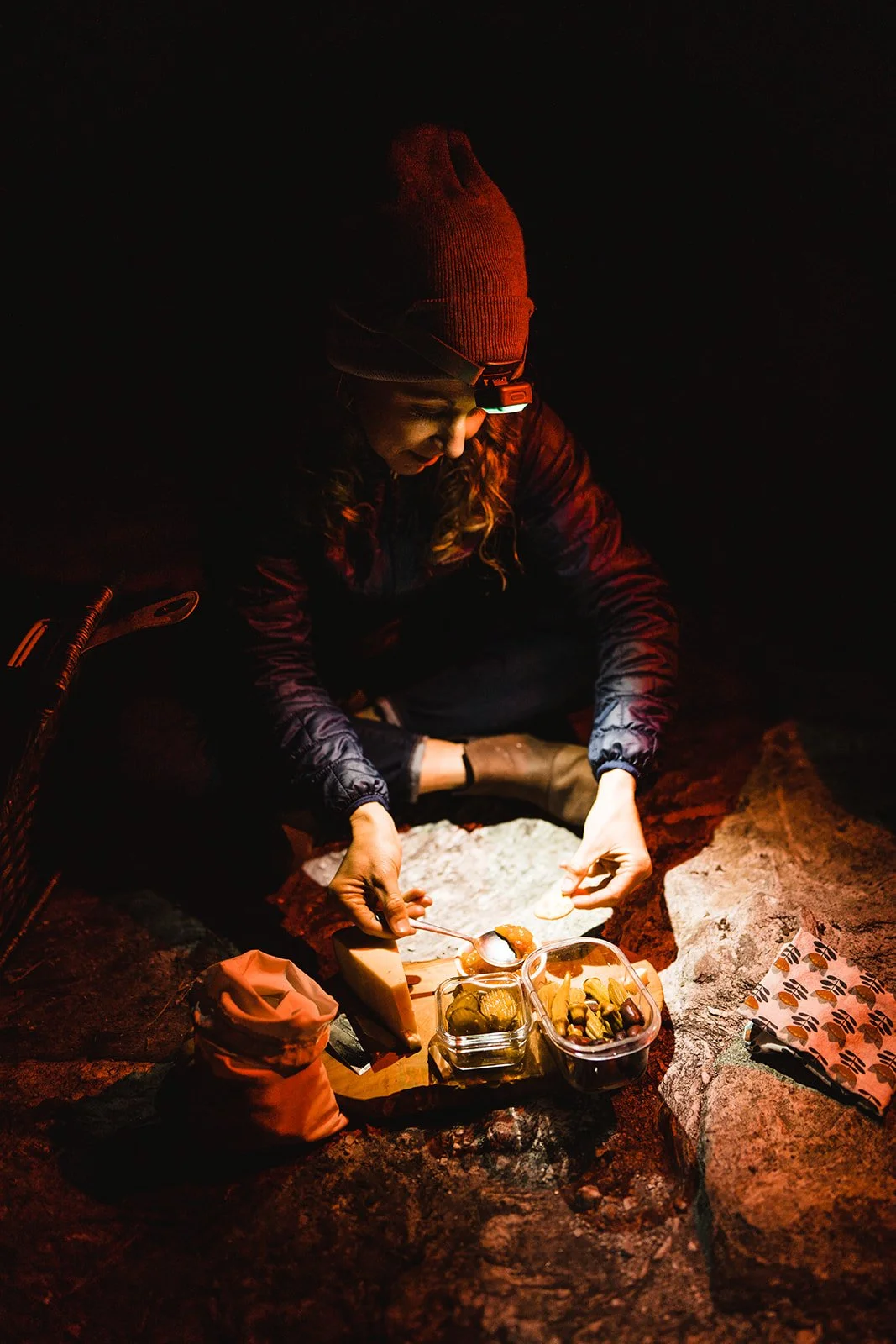 Person wearing a red beanie, night-time outdoor scene, sitting on rocks, preparing food from a snack box with various foods, with a headlamp on, illuminated by a small light