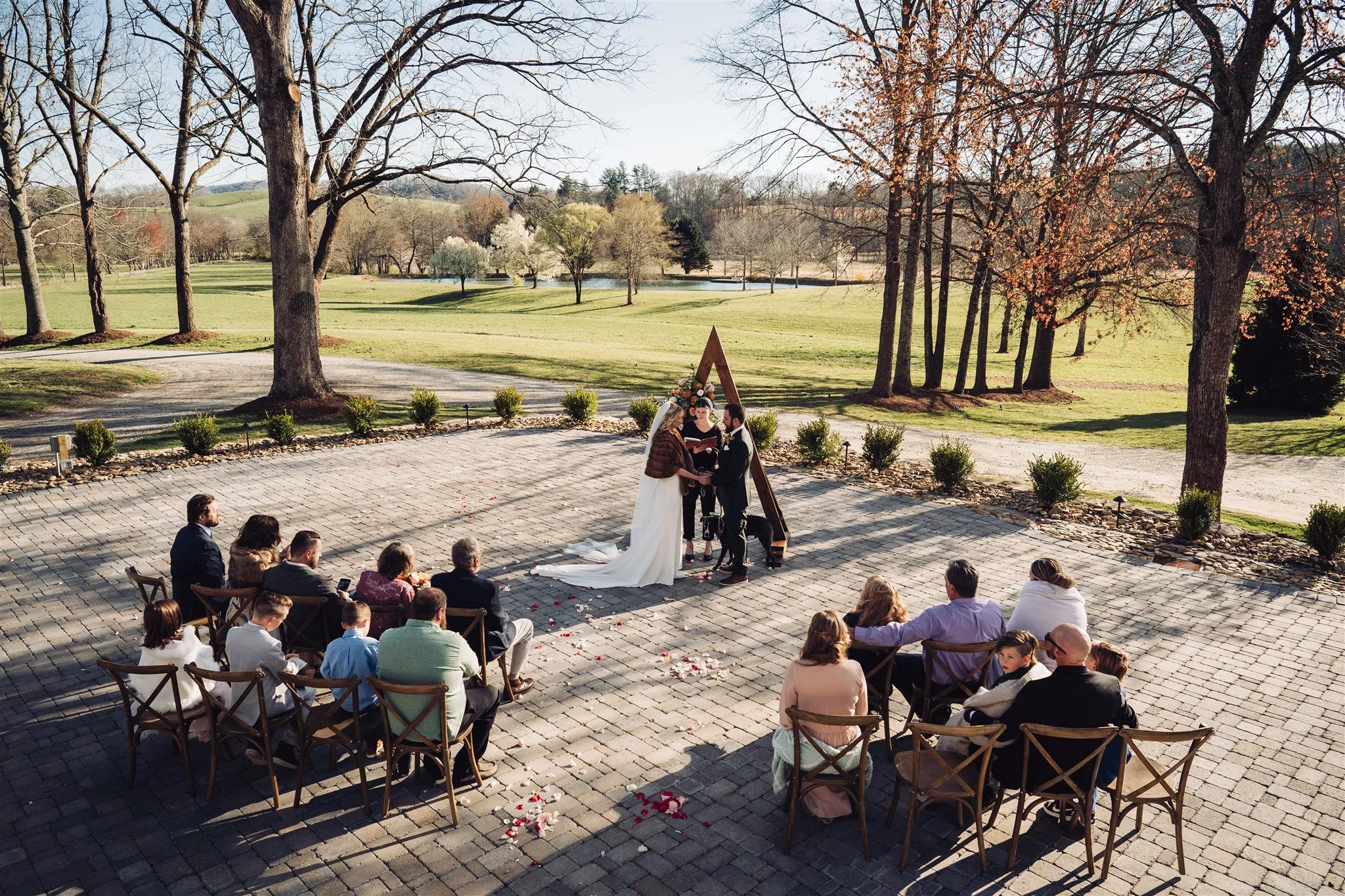 Outdoor wedding ceremony with a bride and groom exchanging vows in front of guests seated on chairs, set on a paved area with trees and a grassy landscape in the background.