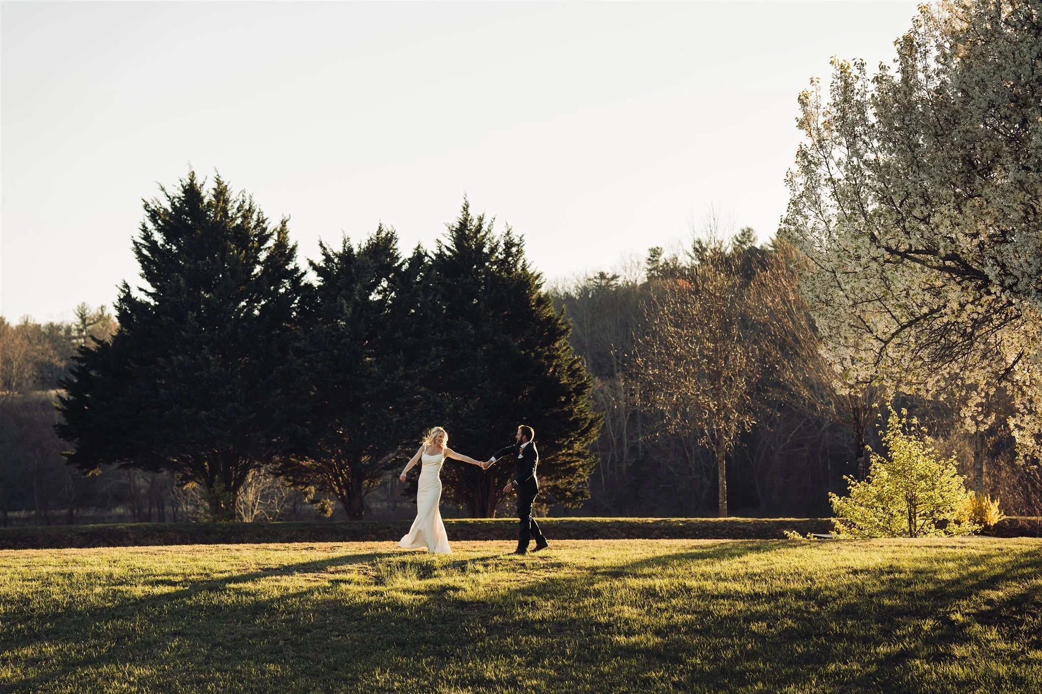 A bride and groom holding hands outdoors during sunset, standing on a grassy field with trees in the background.