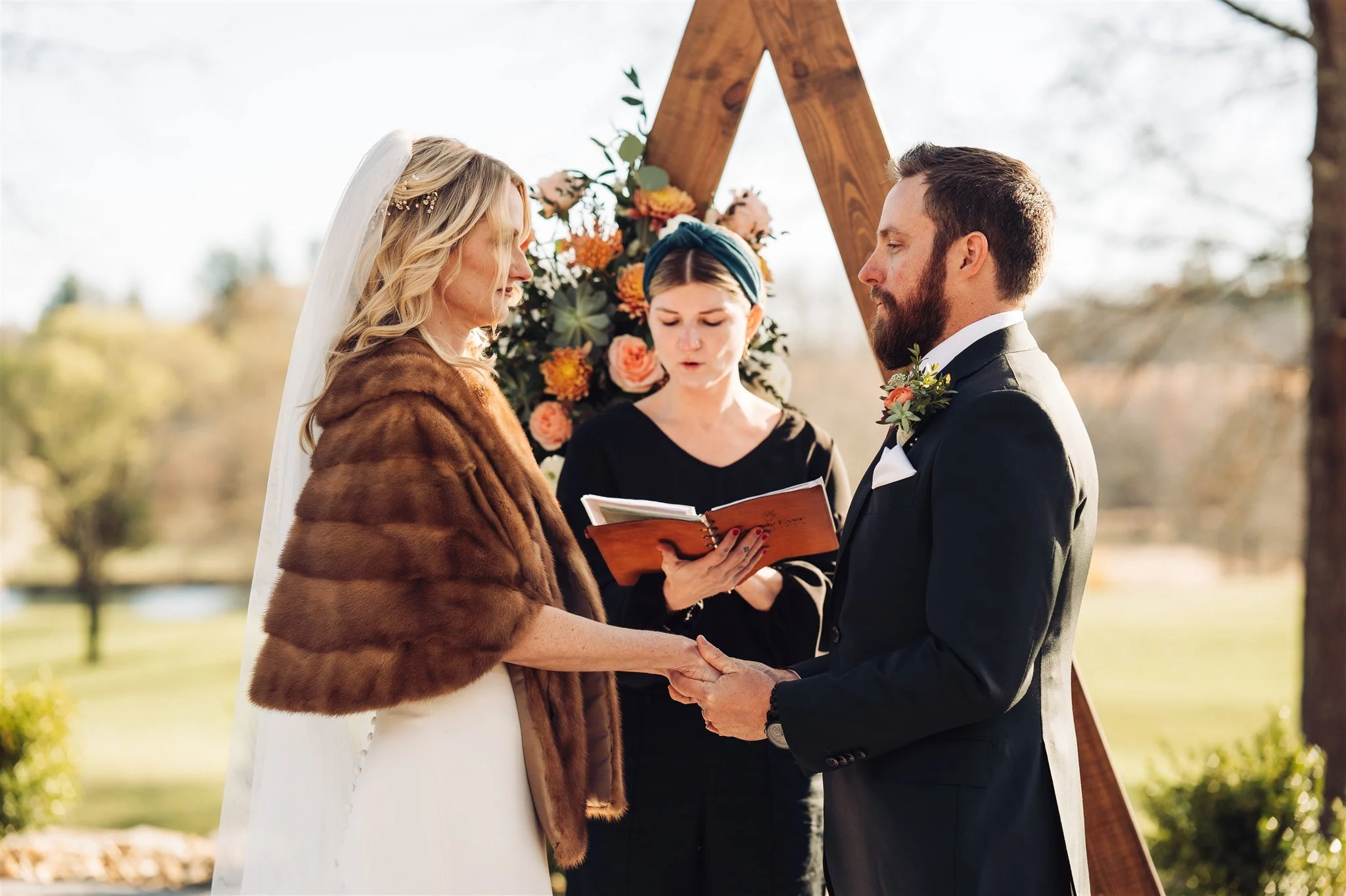 A bride and groom holding hands during an outdoor wedding ceremony, with an officiant reading from a book and a floral arrangement backdrop.