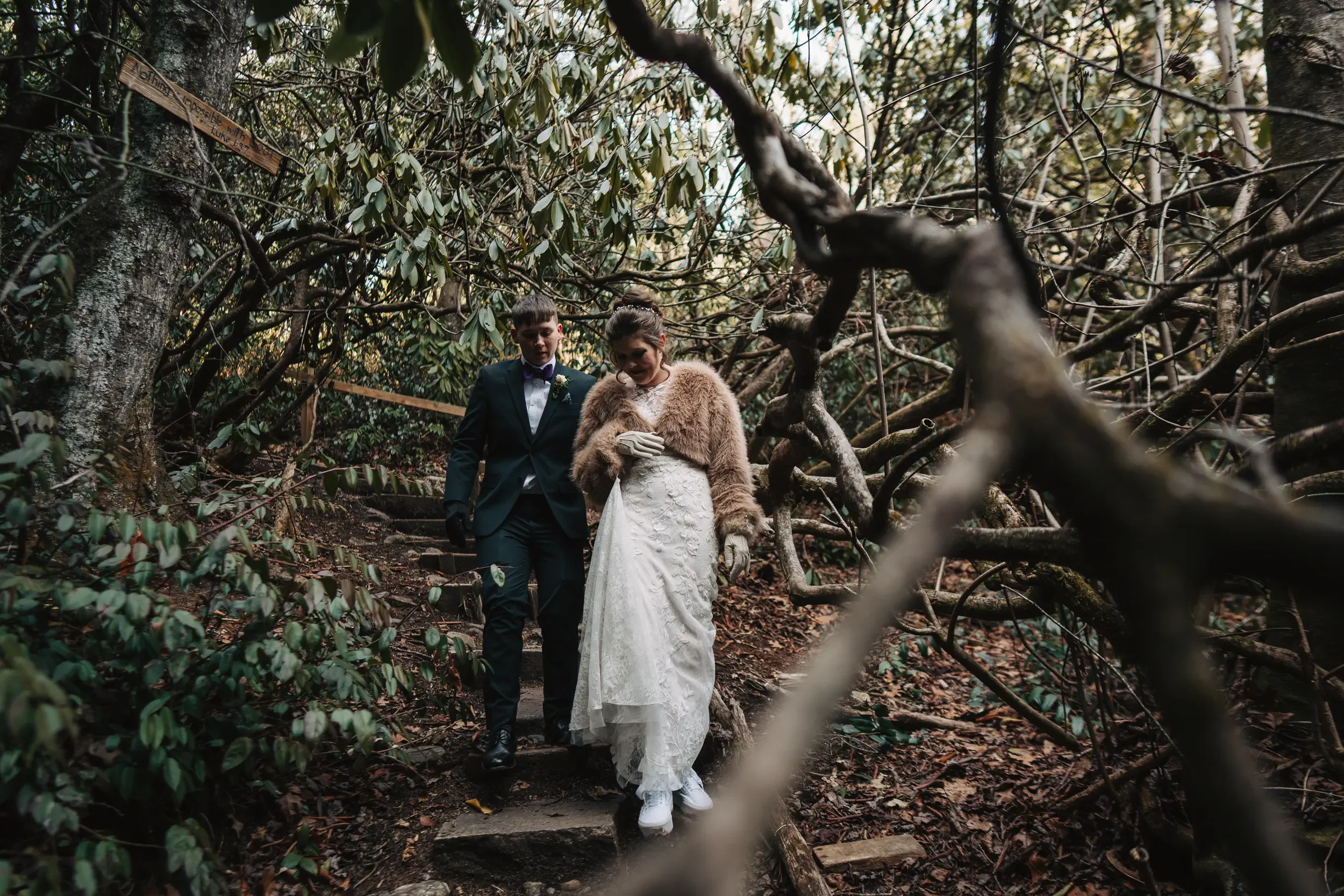 A bride and groom walking through a wooded area during their wedding, with the bride wearing a lace wedding dress and a fur coat, and the groom in a dark suit with a bow tie.