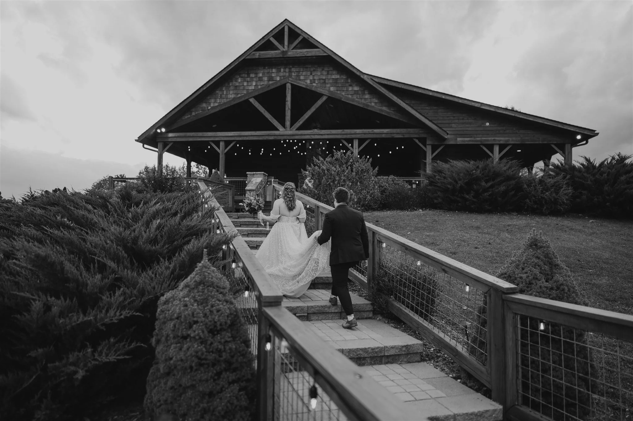 A bride and groom walking up outdoor stairs towards a rustic lodge, holding hands, with string lights overhead and bushes on the sides, in black and white.