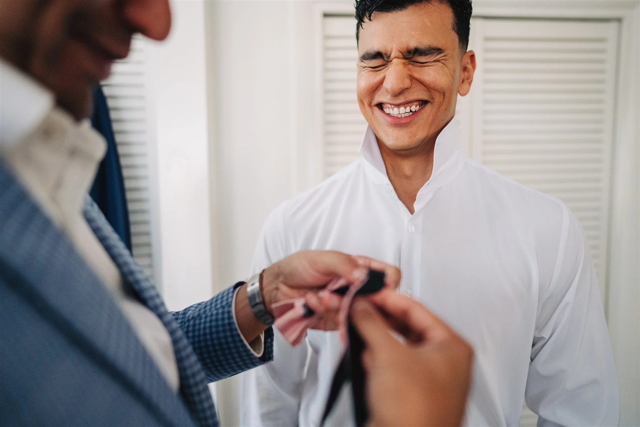 A man in a white shirt laughing while another person is helping him with his attire.