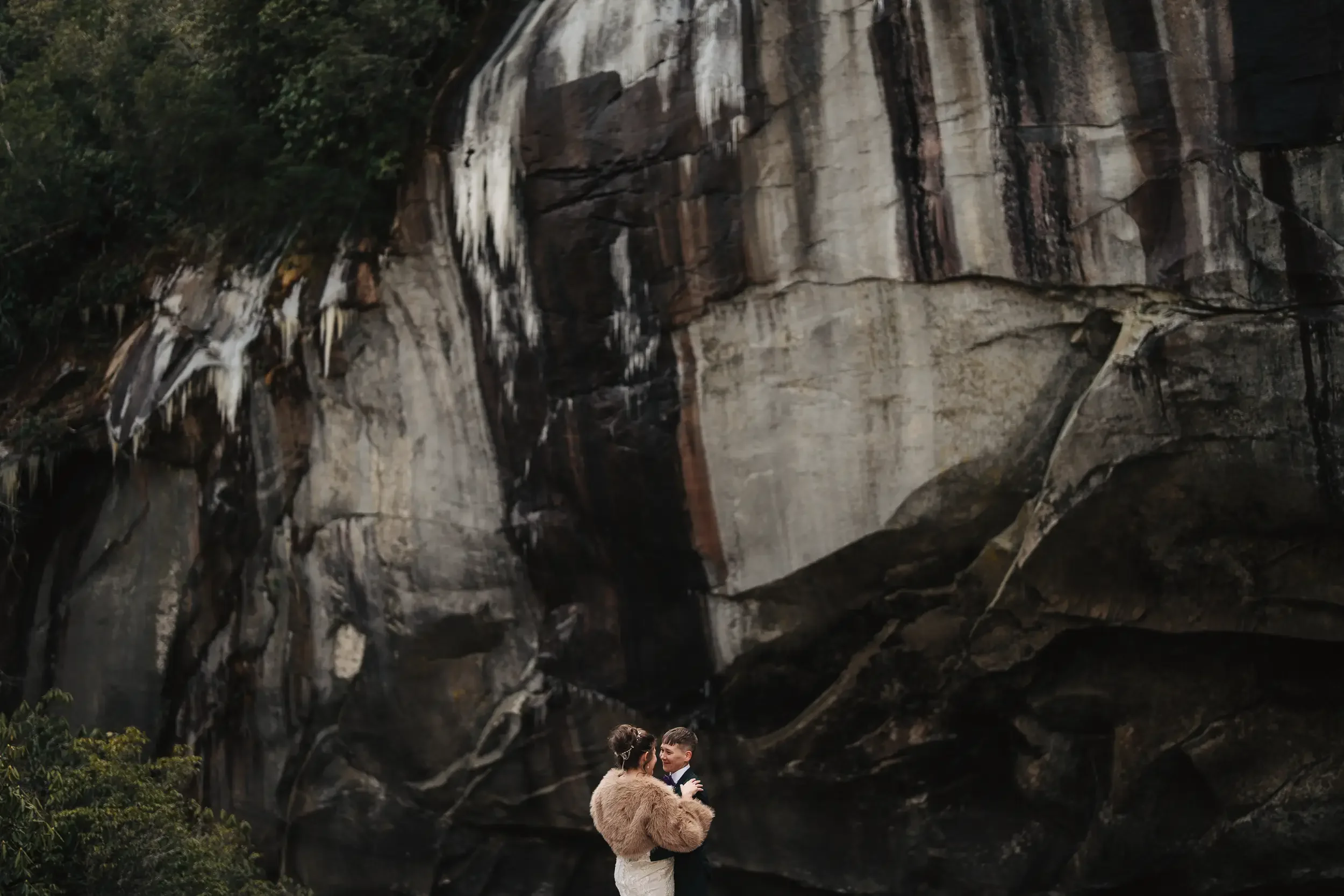 A couple dressed in formal attire, with the woman wearing a fur coat and the man in a suit, standing and embracing in front of a large, natural rock formation with trees and hanging icicles or mineral deposits.