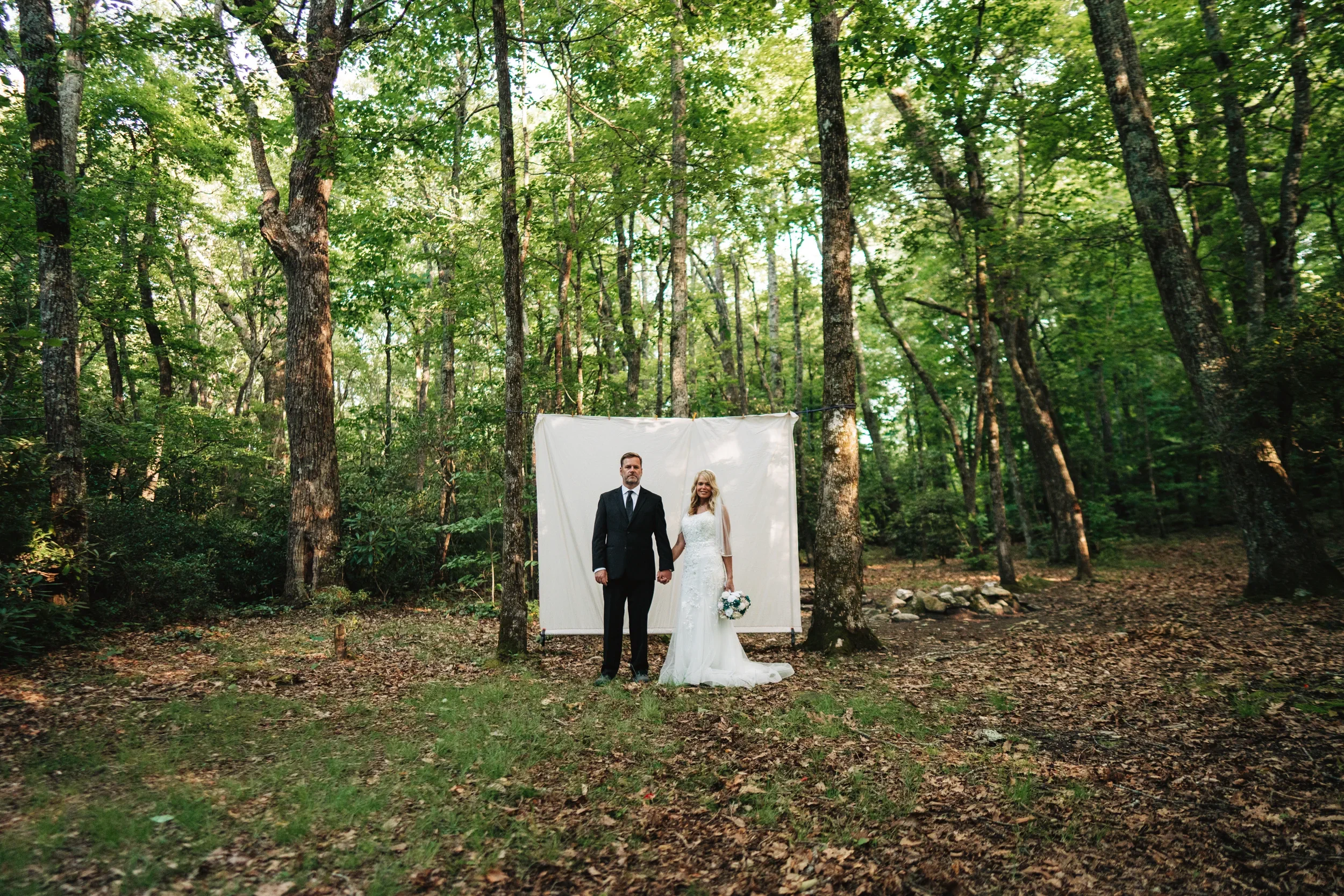 Couple in wedding attire standing hand in hand in a wooded ceremony space, framed by a simple white fabric backdrop among tall trees.