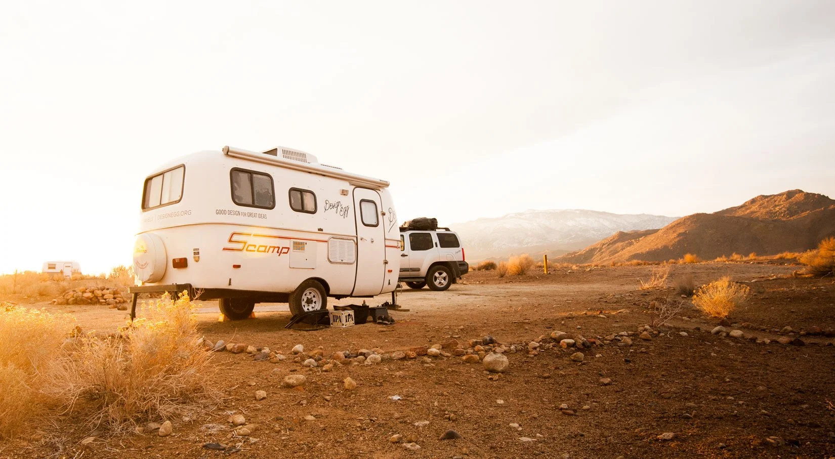 A small white camper trailer named 'Scamp' parked in a desert landscape during sunset, with a black SUV behind it, mountains in the distance, and some sparse desert vegetation and rocks in the foreground.