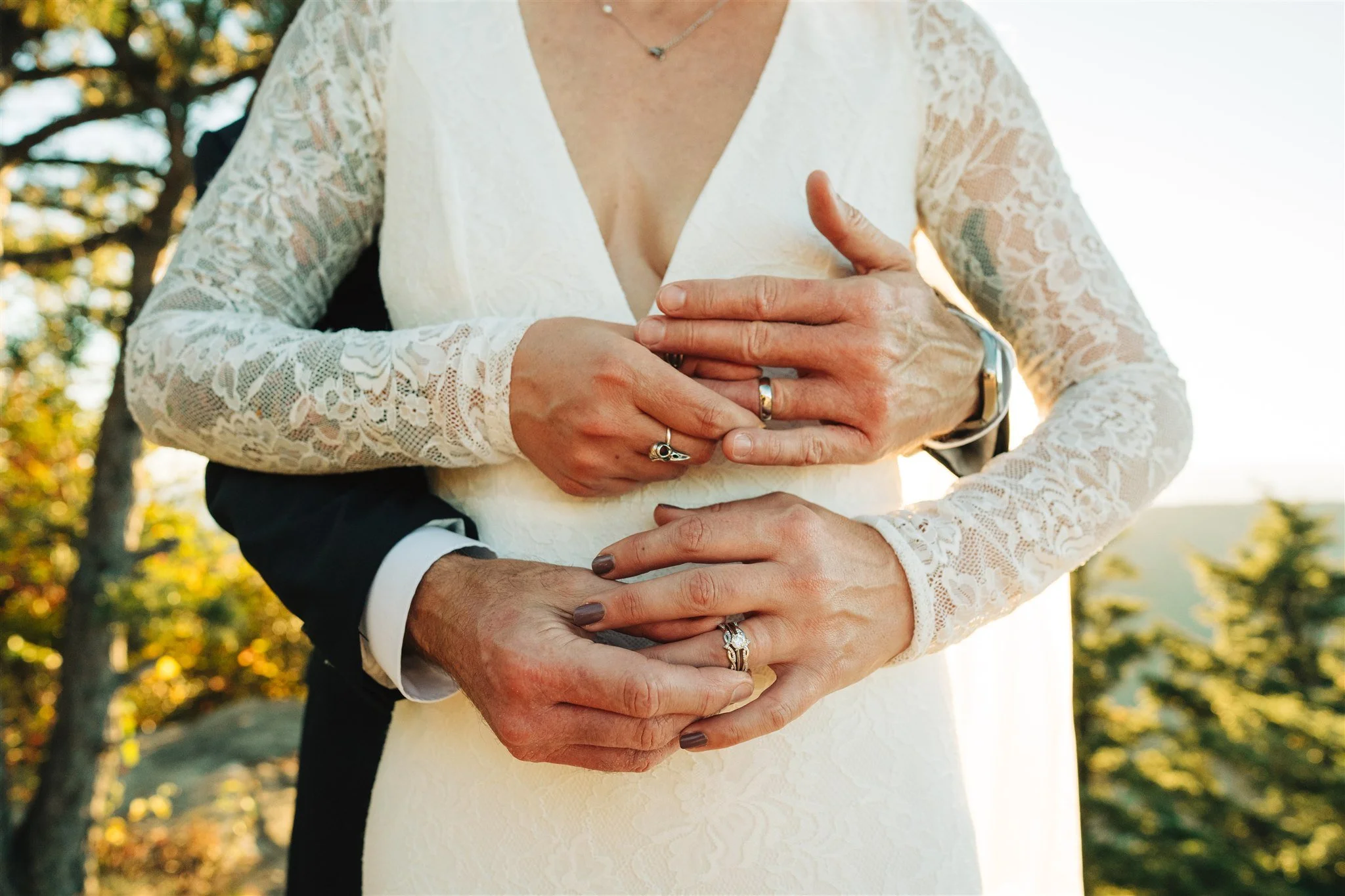 Close-up of a bride in a white lace dress and a groom in a suit, holding hands with wedding rings, outdoors during sunset.