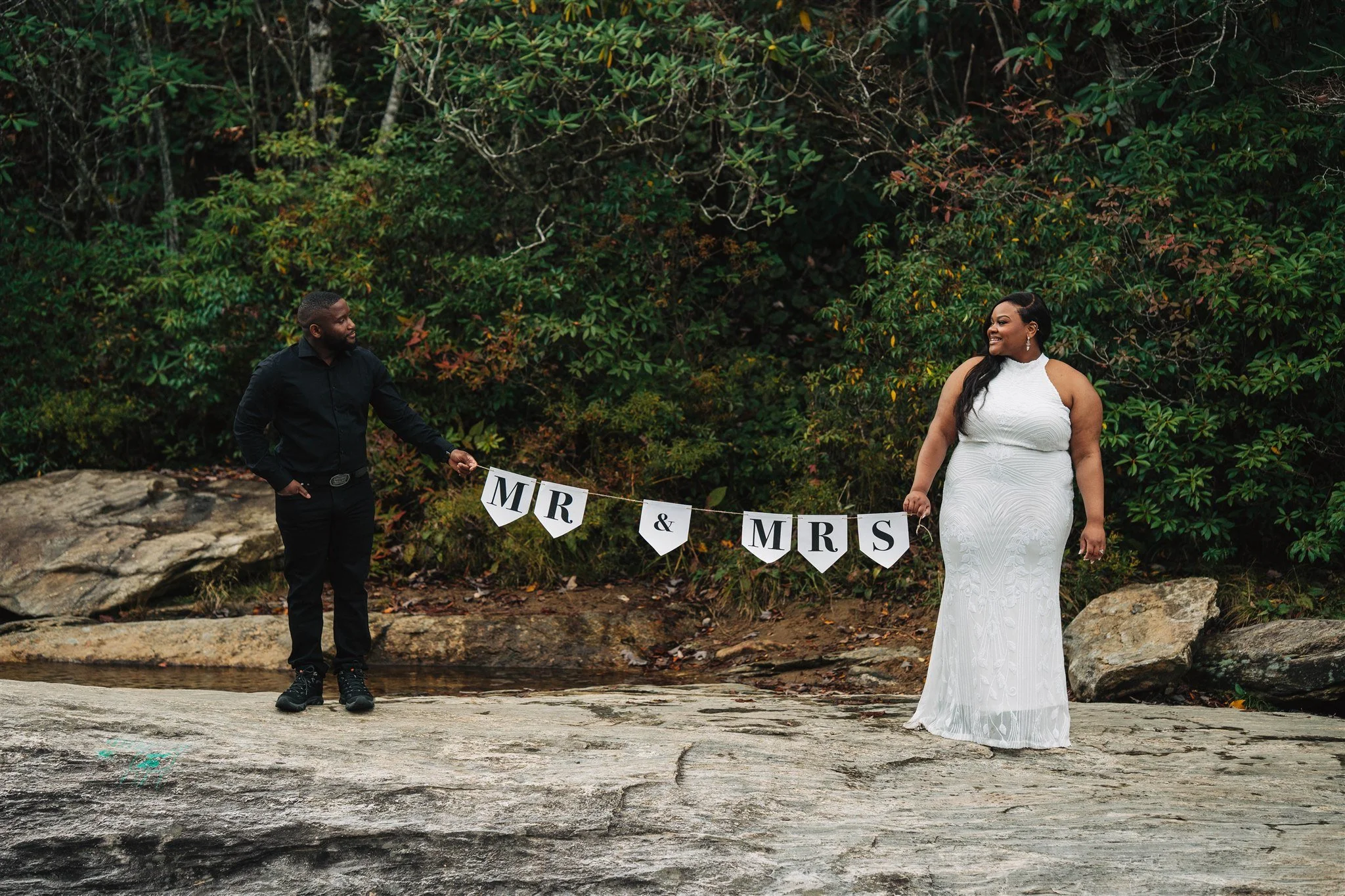 Bride in a white wedding dress and groom holding a sign that reads 'MR & MRS' outdoors with a forest background.
