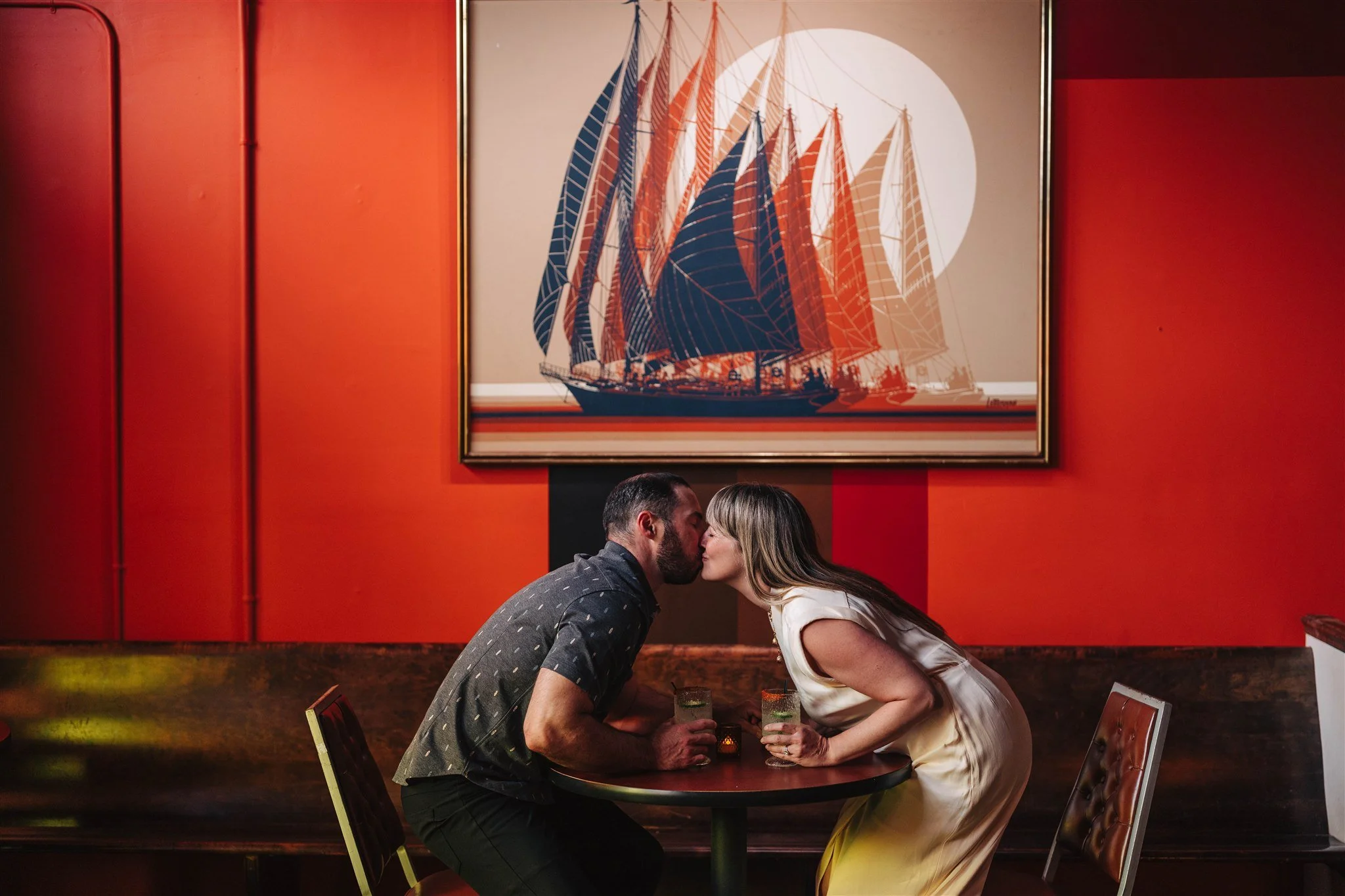 A couple leaning in for a kiss at a bar table with drinks, in front of a large painting of a sailboat with red, blue, and beige sails hanging on a red wall.