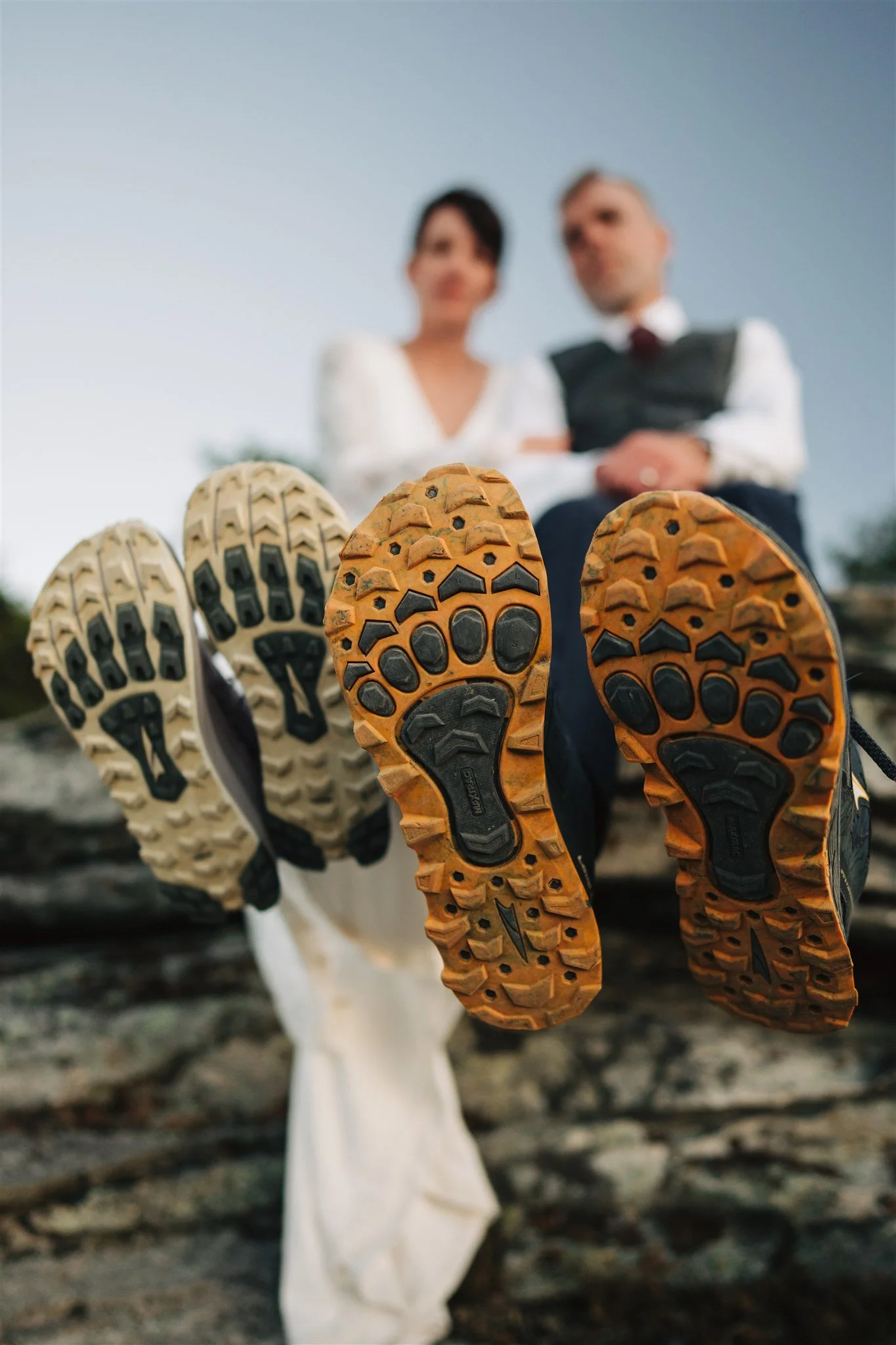 Close-up of the soles of two hiking shoes, with a man and woman dressed in formal attire blurred in the background, sitting on rocks outdoors.