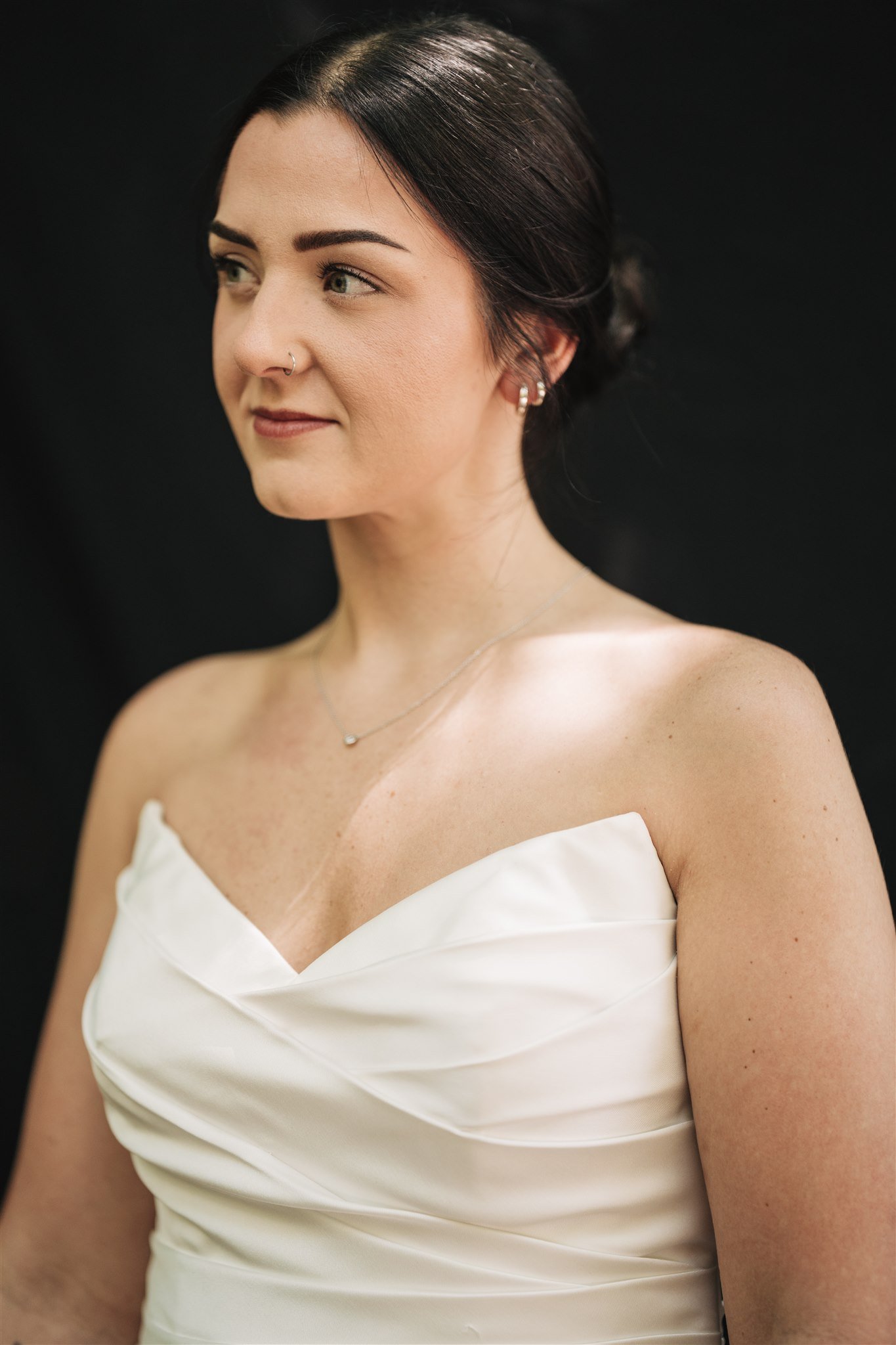 A woman with dark hair styled in an updo, wearing a strapless white dress, small earrings, a nose ring, and a delicate necklace, against a black background.