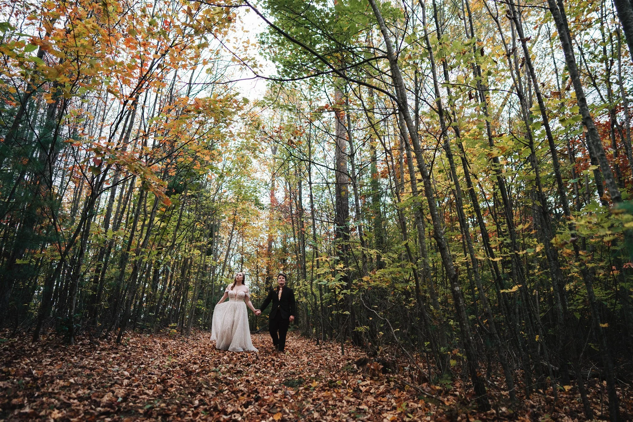 A bride and groom holding hands and walking through a forested area during autumn, surrounded by colorful fall leaves on the ground and trees with orange, yellow, and green foliage.
