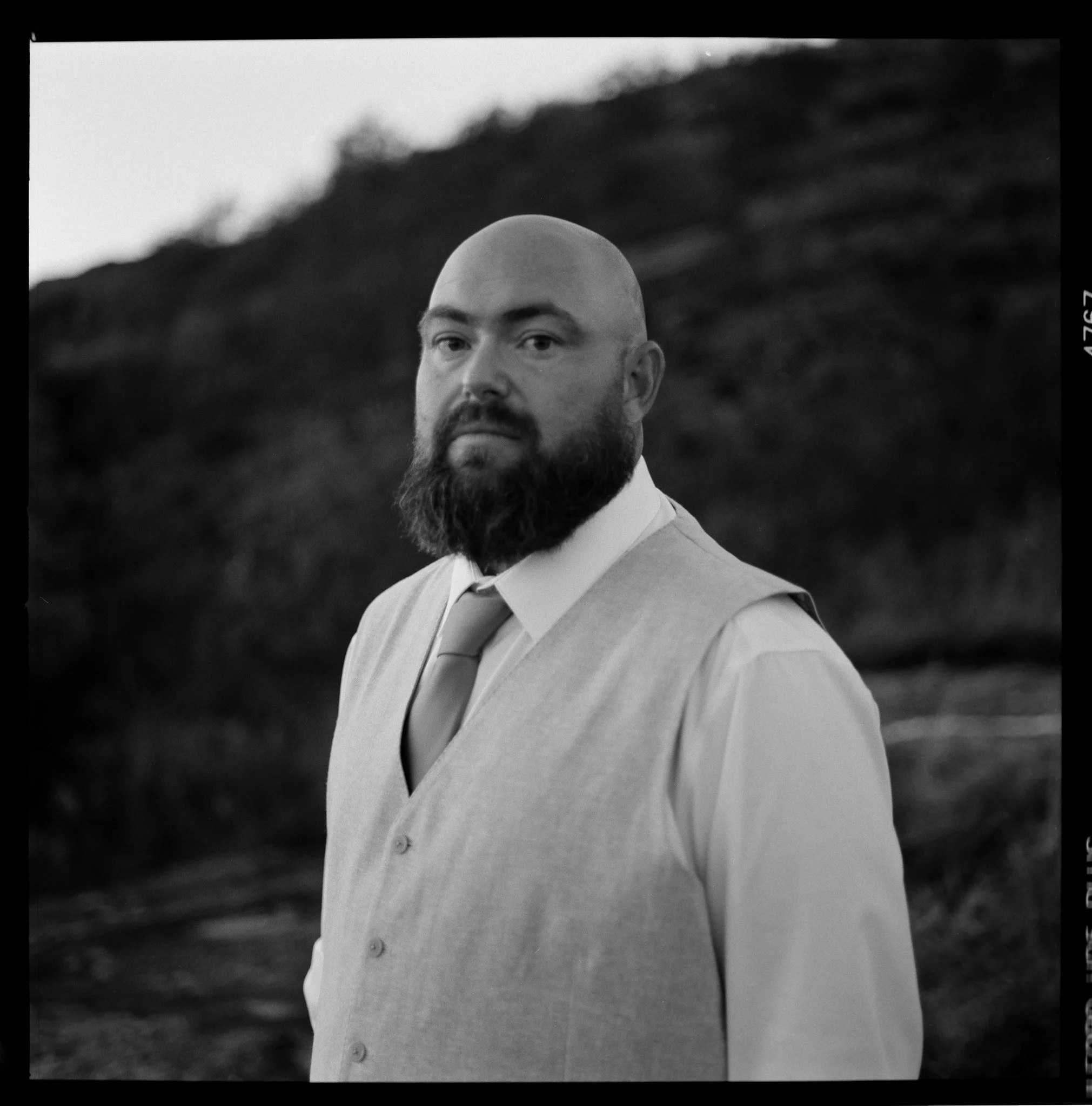 Black and white photo of a man with a beard, wearing a vest and tie, standing outdoors in front of a hillside or mountain.