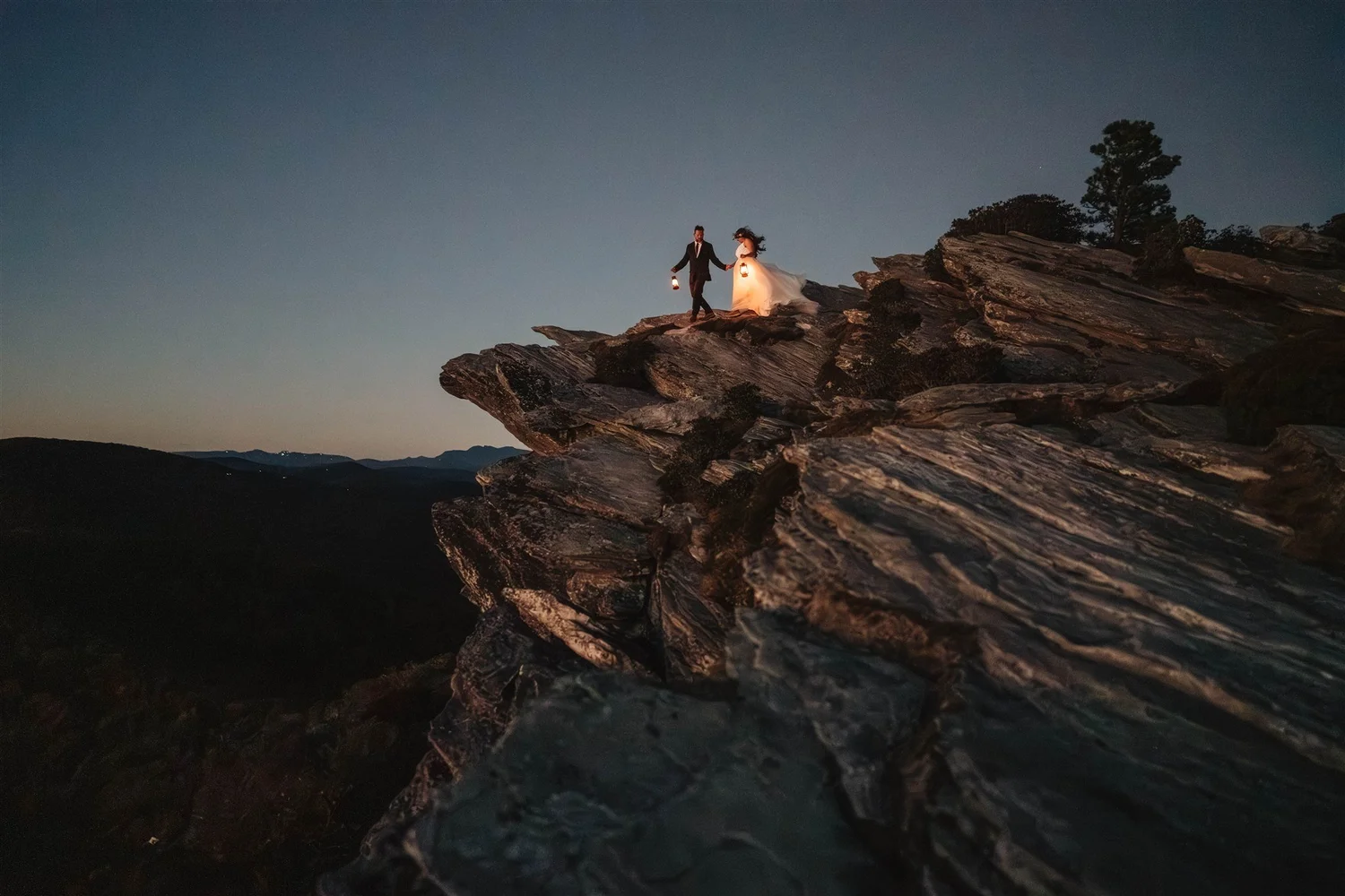 Couple in wedding attire walking on a rocky cliff at dusk with lanterns and mountain views